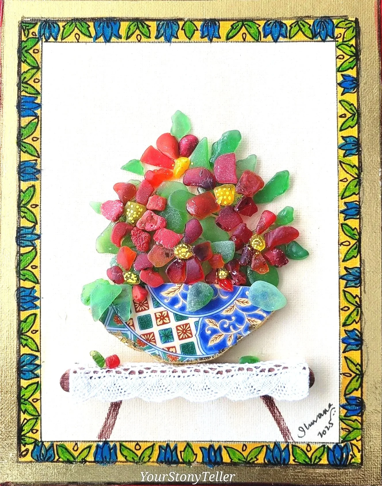 Colorful glass fish-shaped artwork with red, green, purple, and yellow pieces in a blue bowl, set on a small white lace-covered table with a decorated hand-painted border.
