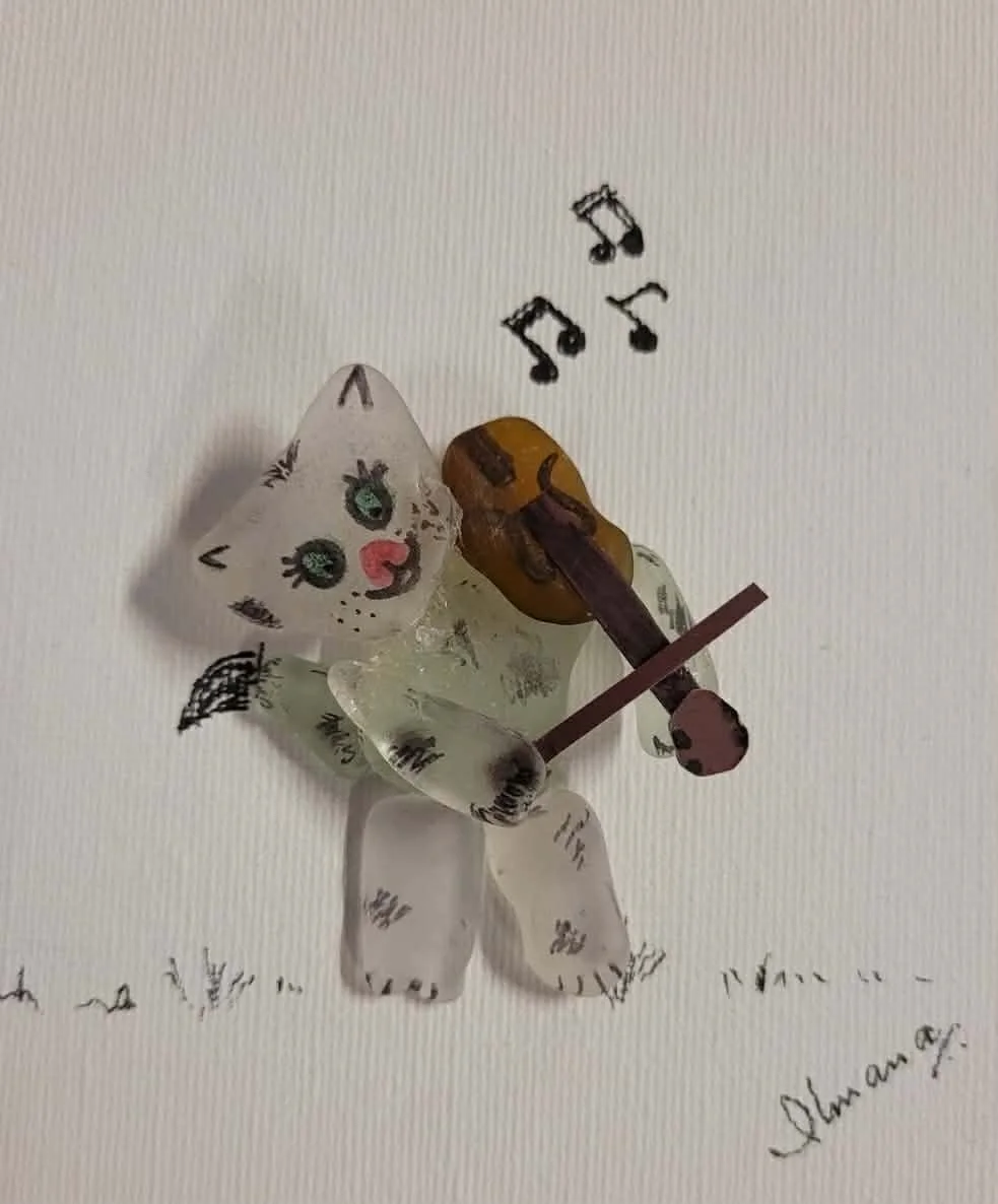 A decorative cat figurine playing a guitar with musical notes above, on a white background with handwritten text made of white and brown seaglass.
