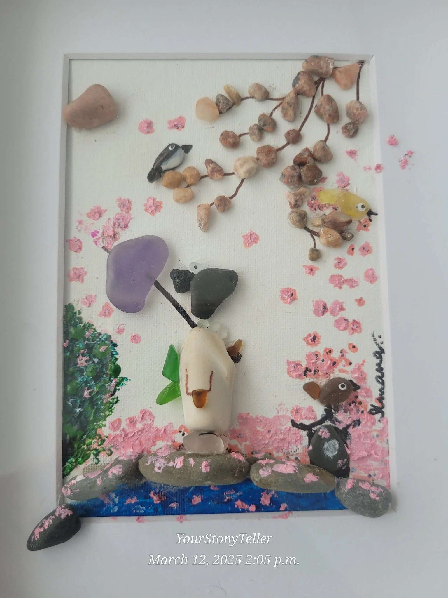 A framed collage of rocks and pebbles arranged to resemble a scene with animals and objects. The scene includes a bird, a squirrel, a cat, and a tree, with pink flower petals and a purple flower, set on a white background with a painted landscape. The frame has a few larger rocks at the bottom, and a signature or name on the right side.