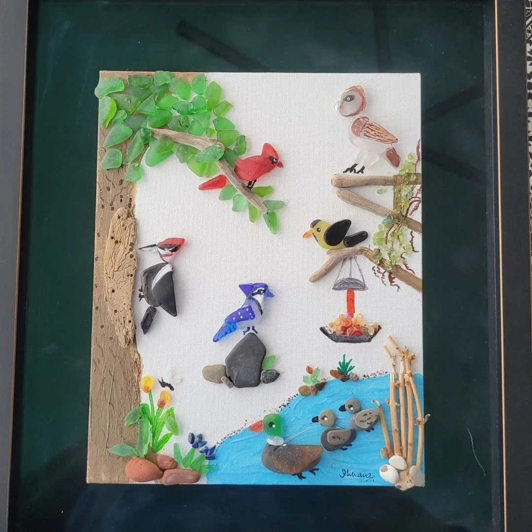 Sea-themed craft scene with colorful stones and driftwood resembling birds, trees, and water on a white background.