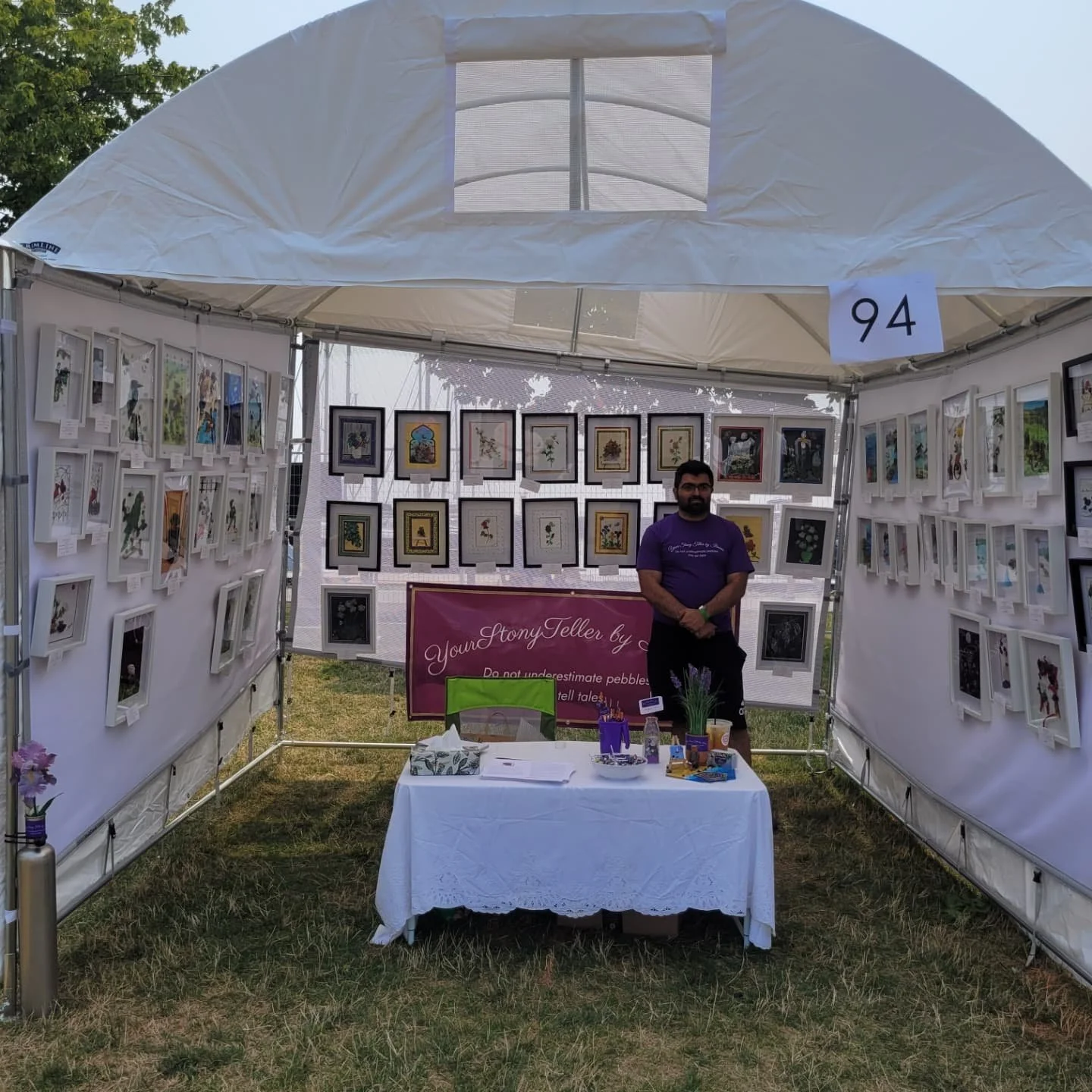 An outdoor art booth with framed artwork displayed on white panels, a table with art supplies and a sign that reads 'YourStoryTeller by C' with a slogan, a man standing behind the table, and a numbered sign '94' hanging from the tent