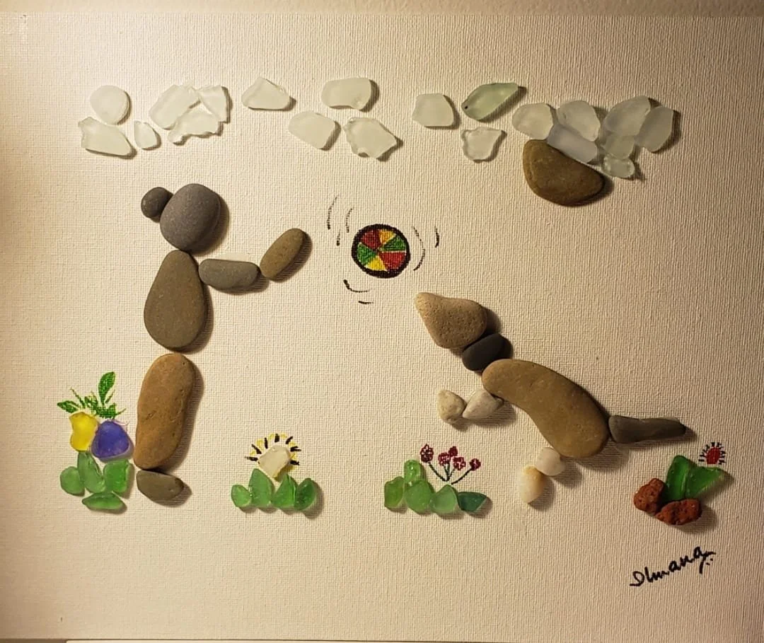 Artwork made of arranged rocks and pebbles on a surface, depicting a scene with a person holding a sphere, plants, flowers, clouds, and handwritten signature.
