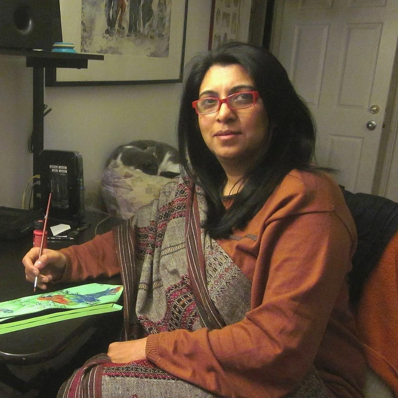 A woman with black hair and red glasses sitting at a desk, holding a paintbrush, with a painting of a fish in front of her.