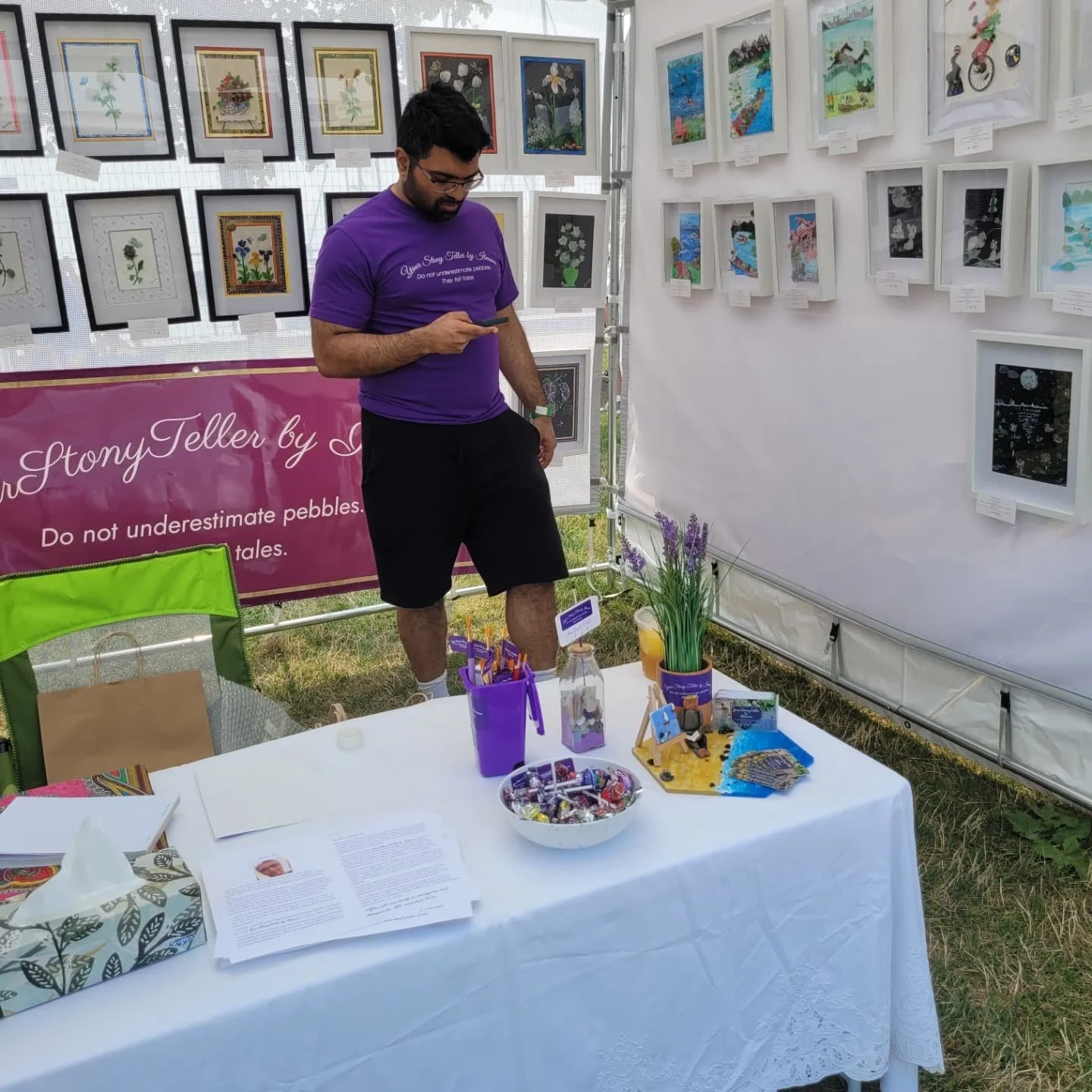 A man in a purple t-shirt and black shorts standing at an art booth, looking at his phone. The booth displays framed artwork on the walls behind him and various items on a white table in front. The background shows outdoor grass and a pink banner with white text.