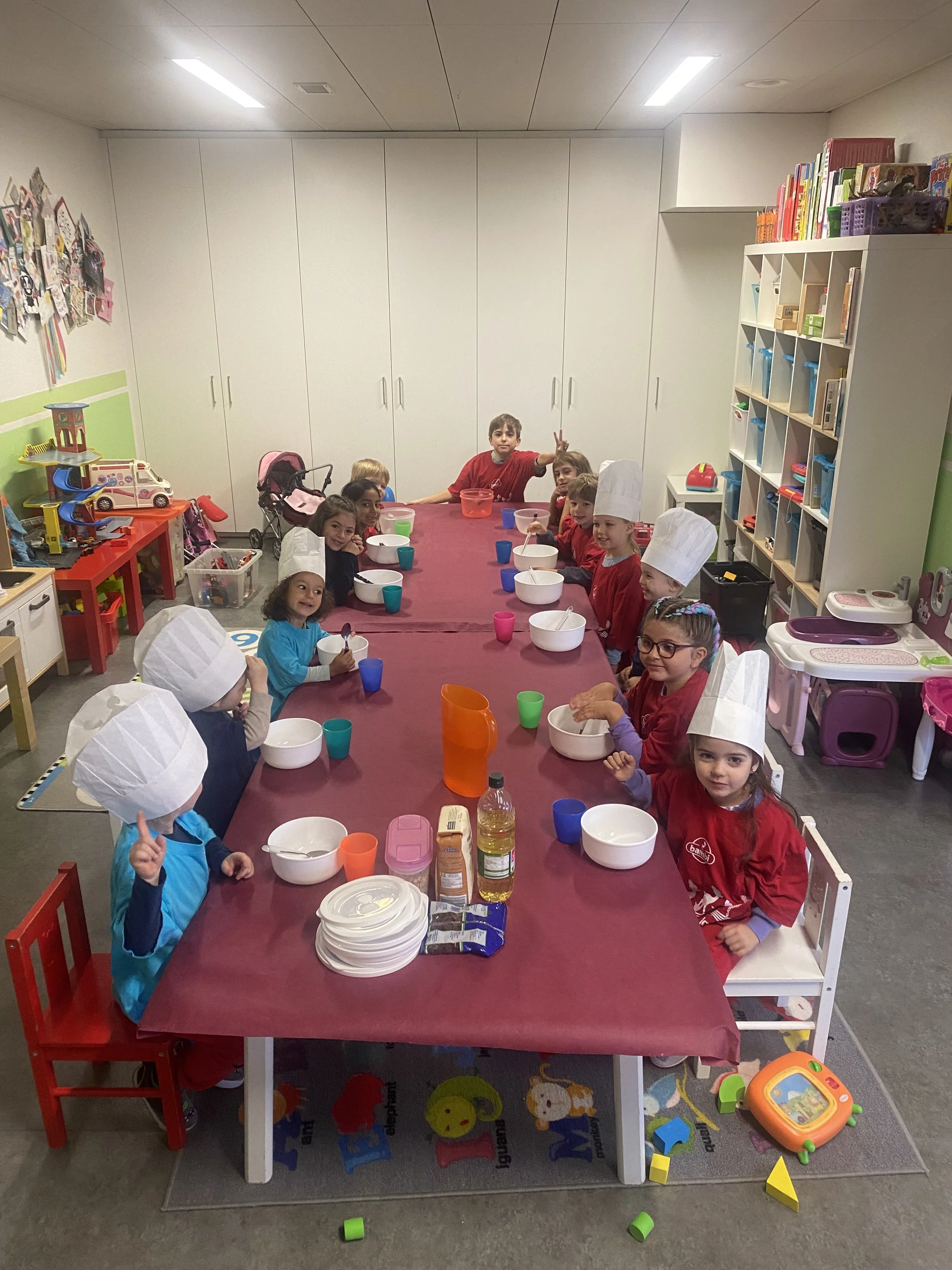 Kinder in einer Kindertagesstätte beim Kochen oder Backen, tragen Kochmützen, sitzen an einem langen Tisch mit Schüsseln und Zubehör.