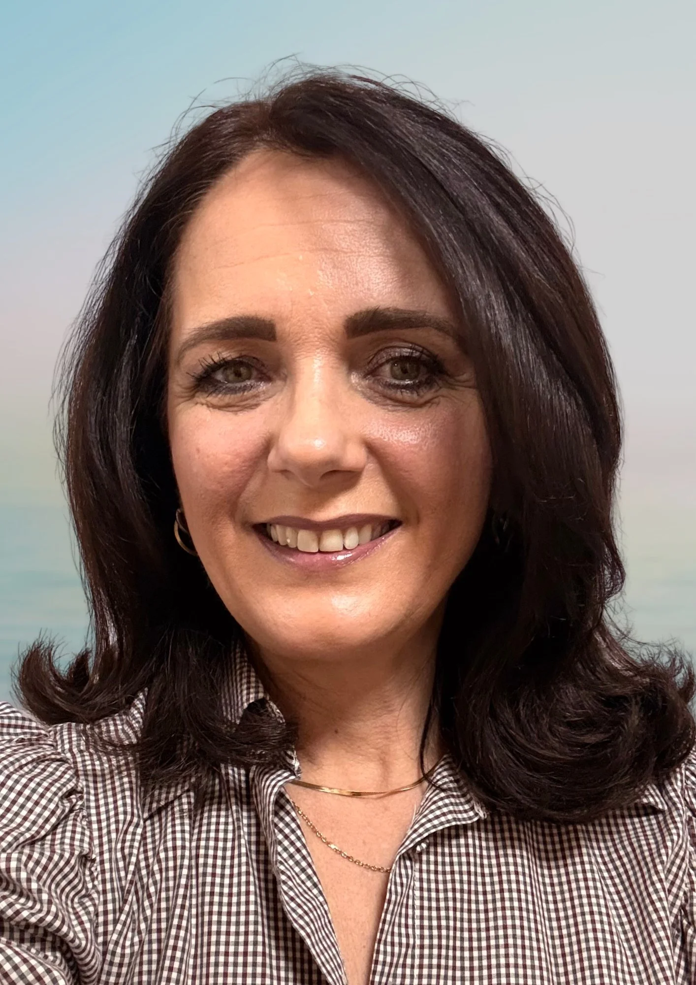 Porträt einer Frau mit braunen Haaren, dunklen Augen, grauem, kariertem Hemd und Schmuck, vor einem unscharfen natürlichen Hintergrund.