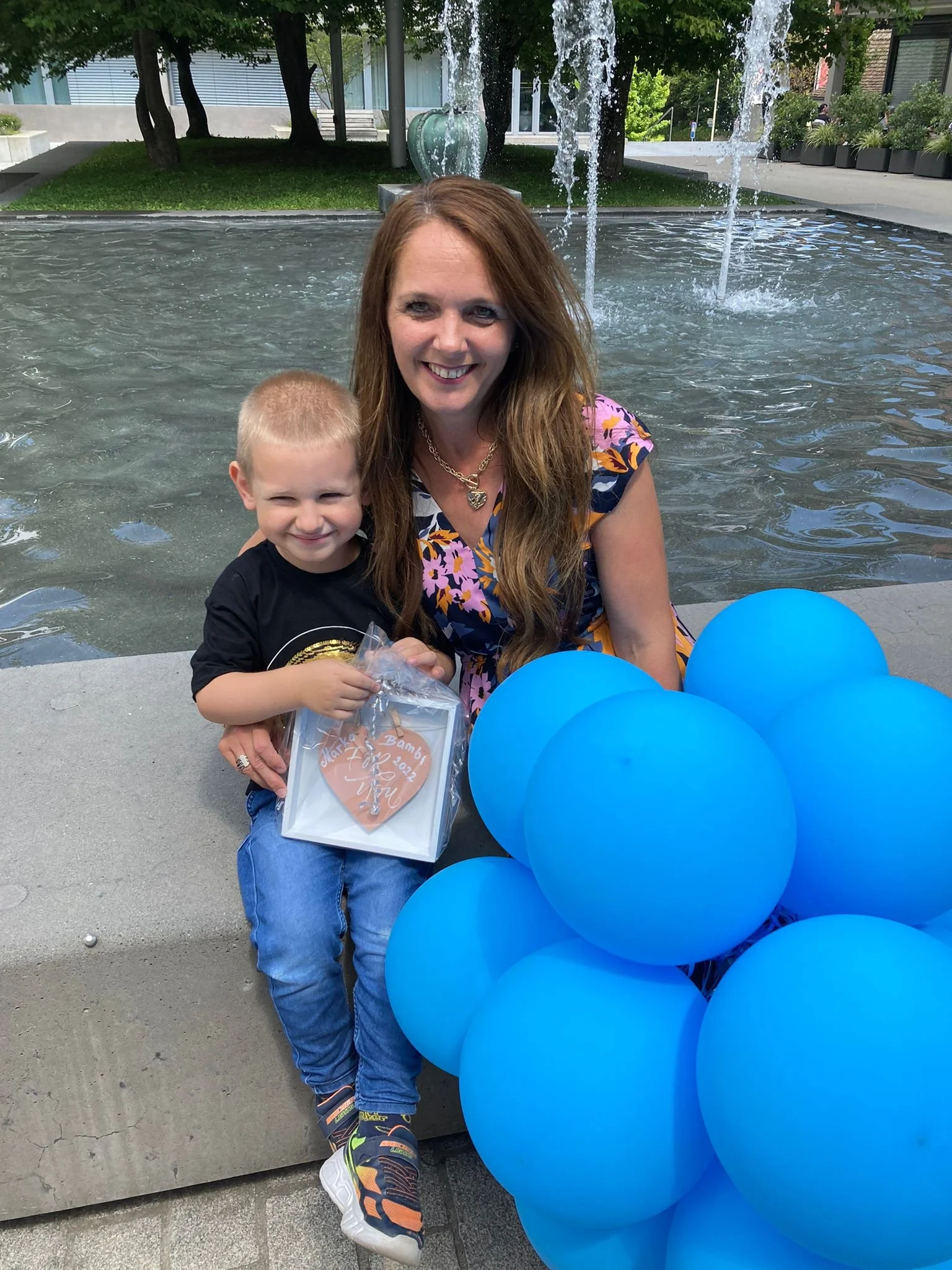 Eine Frau und ein Kind sitzen an einem Brunnen im Freien, das Kind hält ein Geschenk in der Hand, vor ihnen eine Gruppe blauer Ballons, im Hintergrund Bäume und moderne Gebäude.