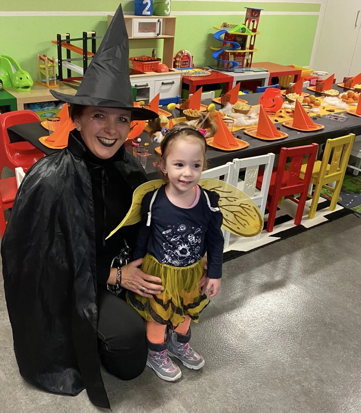 Ein Erwachsener und ein Kind in Halloween-Kostümen stehen vor einer festlich dekorierten Tafel, die mit orangefarbenen Papptellern, Trinkgläsern und Dekoartikeln für eine Halloween-Party gedeckt ist.