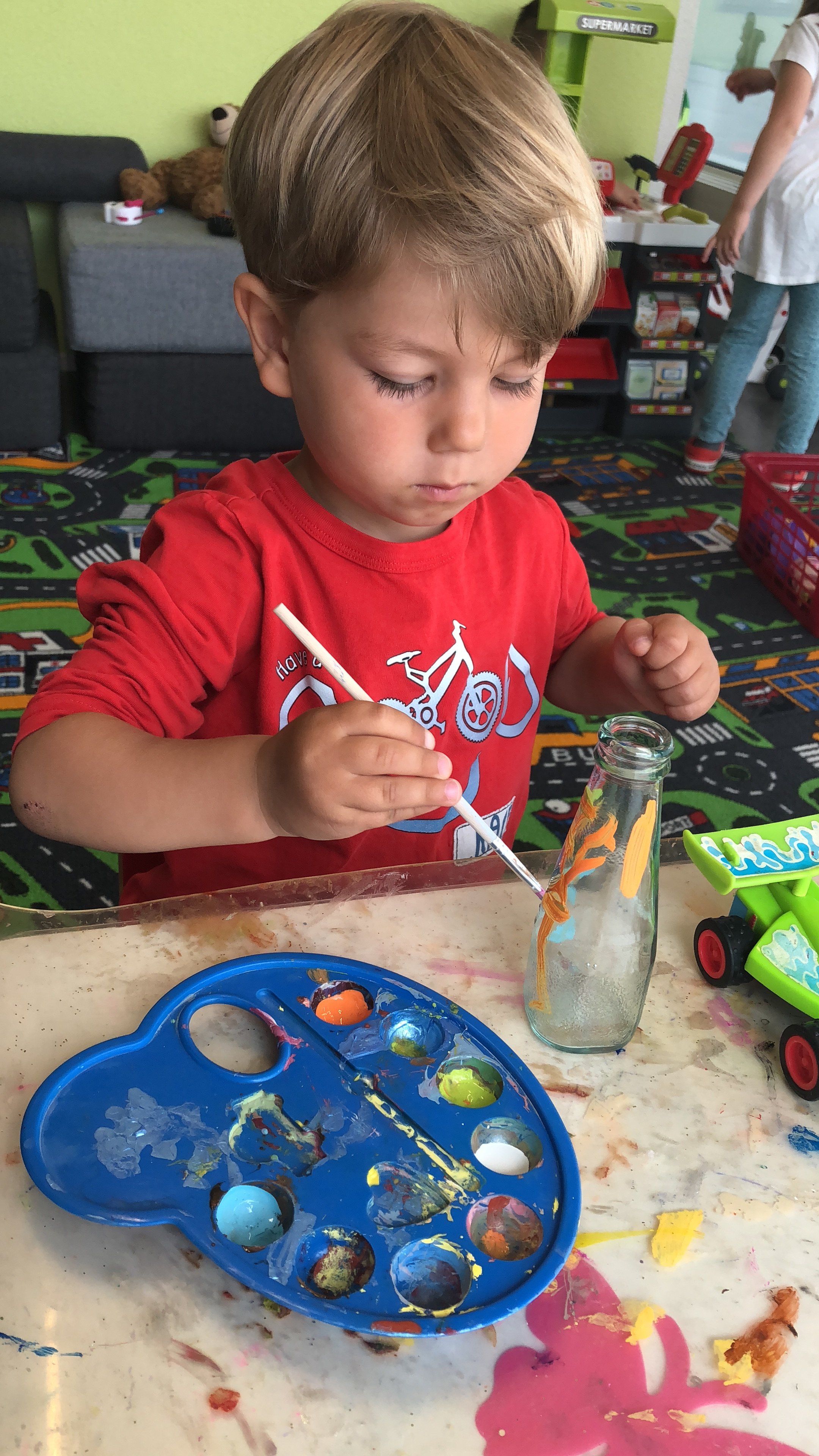 Ein Junge in einem roten T-Shirt, der mit Pinteln arbeitet und eine leere Glasflasche bemalt. Der Tisch ist mit Farbe überspritzt, und im Hintergrund sind Spielzeug und eine Spielzeuggeschäft zu sehen.