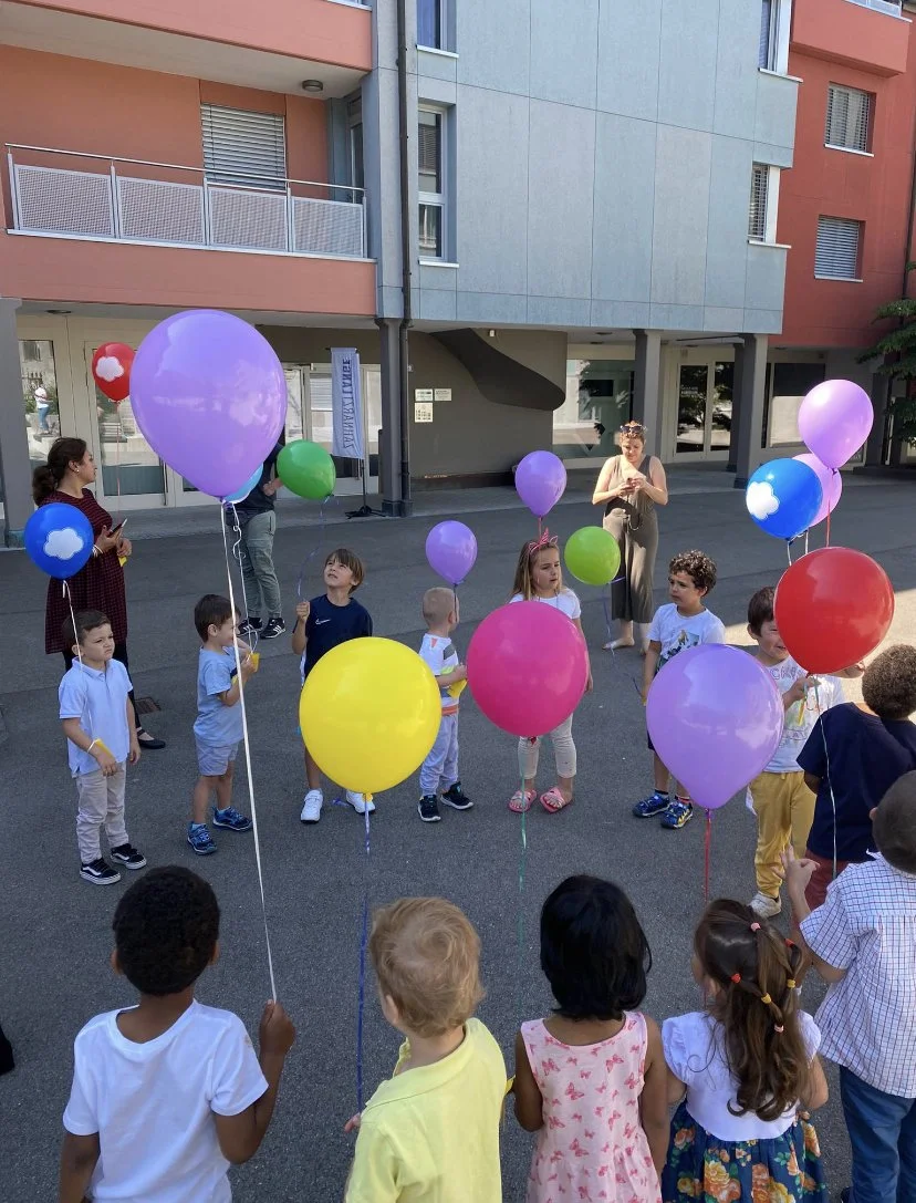 Gruppe von Kindern mit bunten Luftballons, die draußen vor einem modernen Gebäude stehen, mit einer erwachsenen Begleitperson im Hintergrund.
