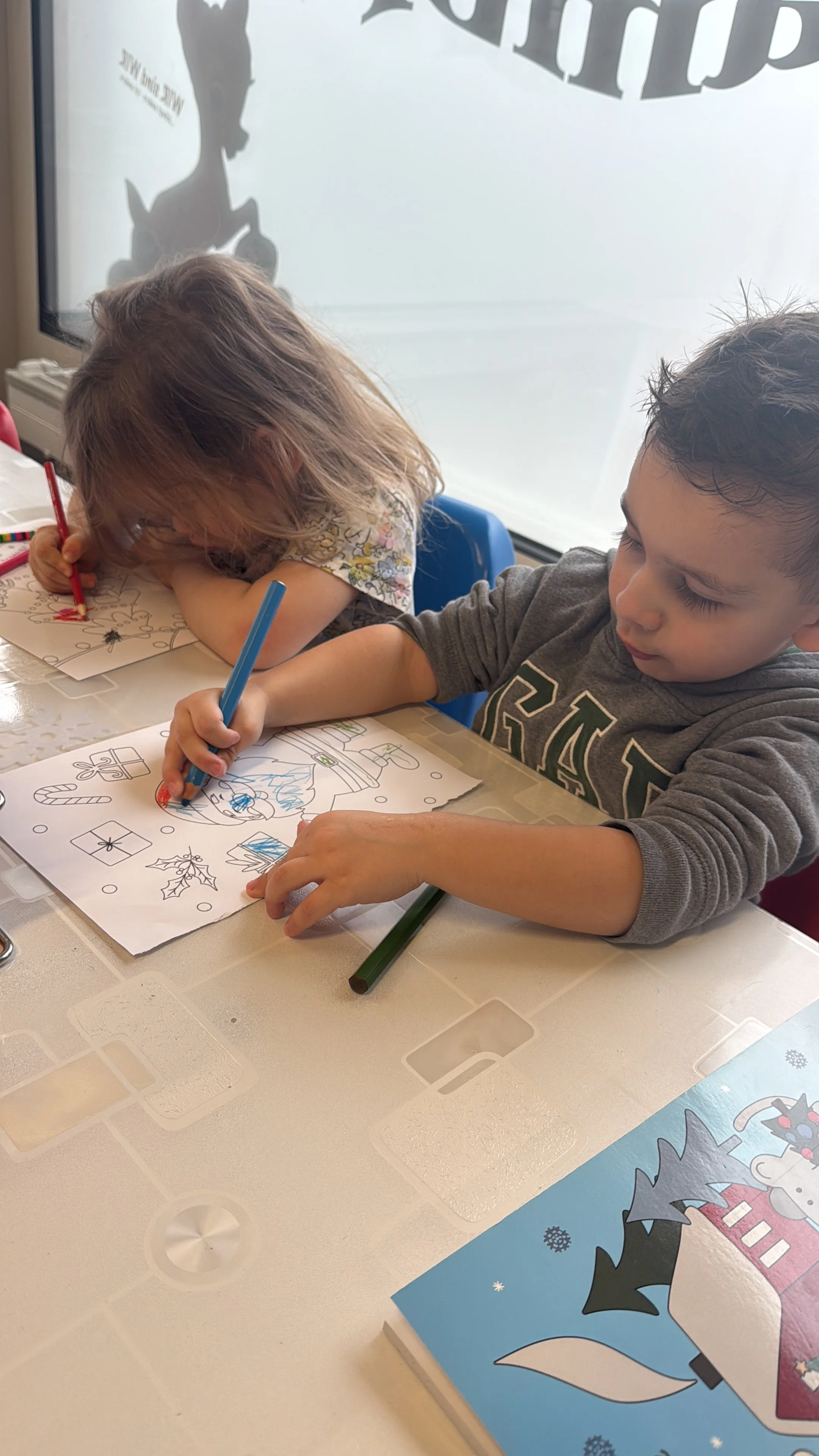Zwei Kinder sitzen an einem Tisch und malen Weihnachtsbilder aus, eines mit grünen und blauen Farben, das andere mit roter Farbe.