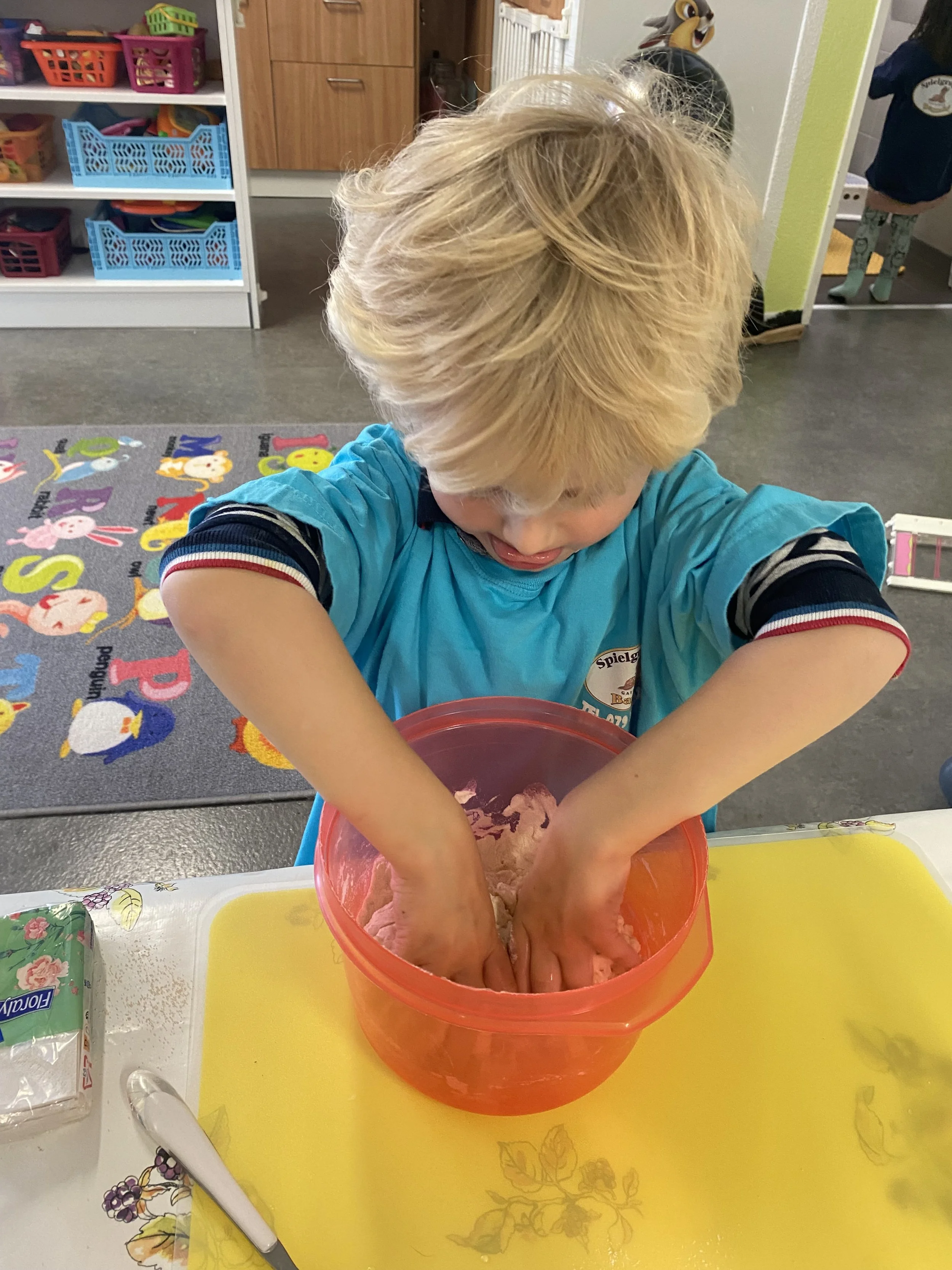 Ein Kinderjunge mit blonder Haar, der mit den Händen in einer roten Schüssel mit Teig arbeitet, auf einem weißen Tisch mit einem gelben Schneidebrett.