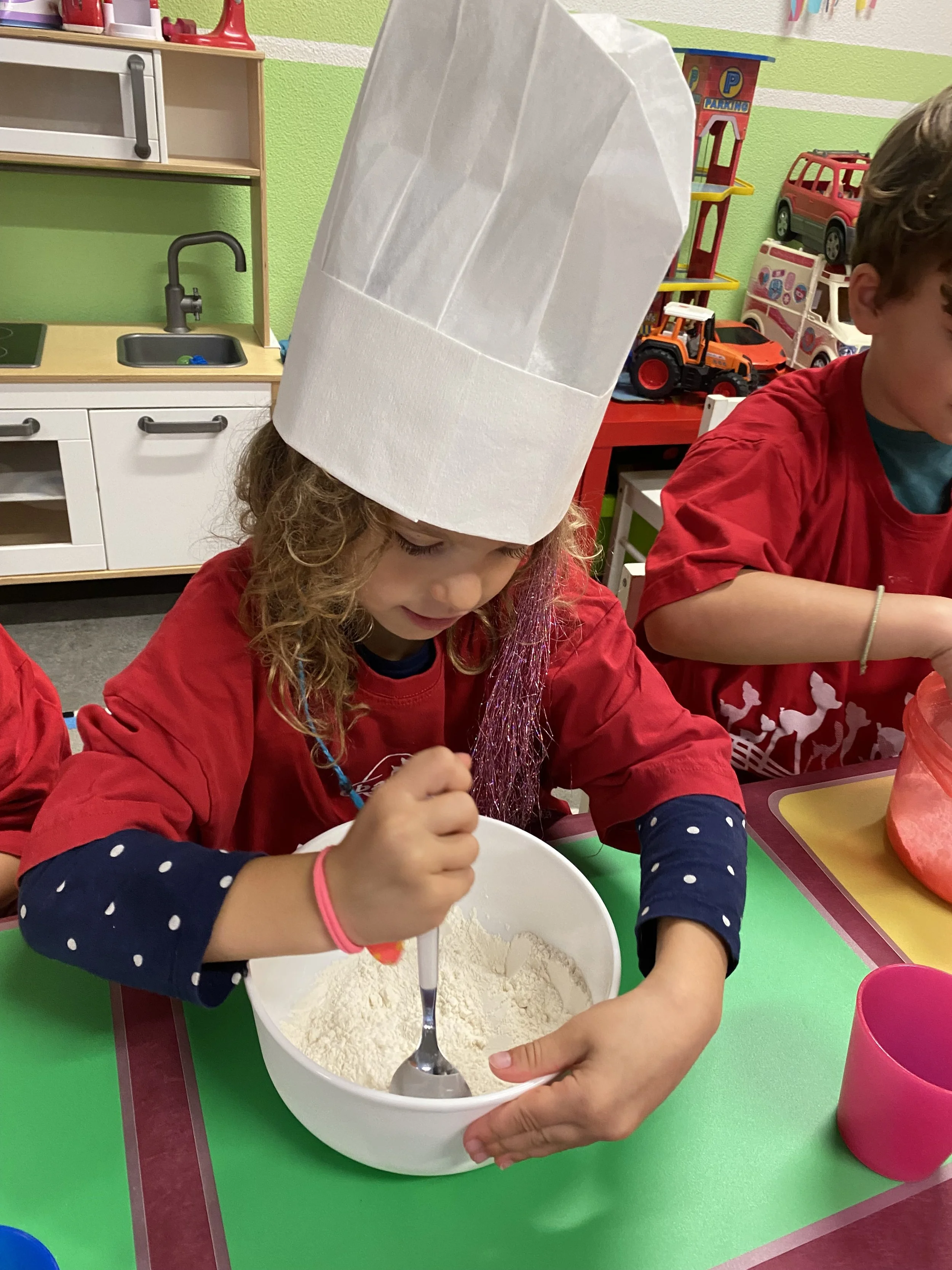 Ein Mädchen trägt einen weißen Kochmütze, rührt in einer weißen Schüssel mit Mehl beim Backen in einer bunten, kindgerechten Spielküche.