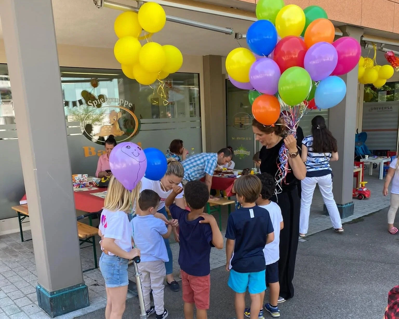 Kinder feiern Geburtstag im Freien mit bunten Luftballons und Spieltischen, Erwachsene beteiligen sich an den Aktivitäten.