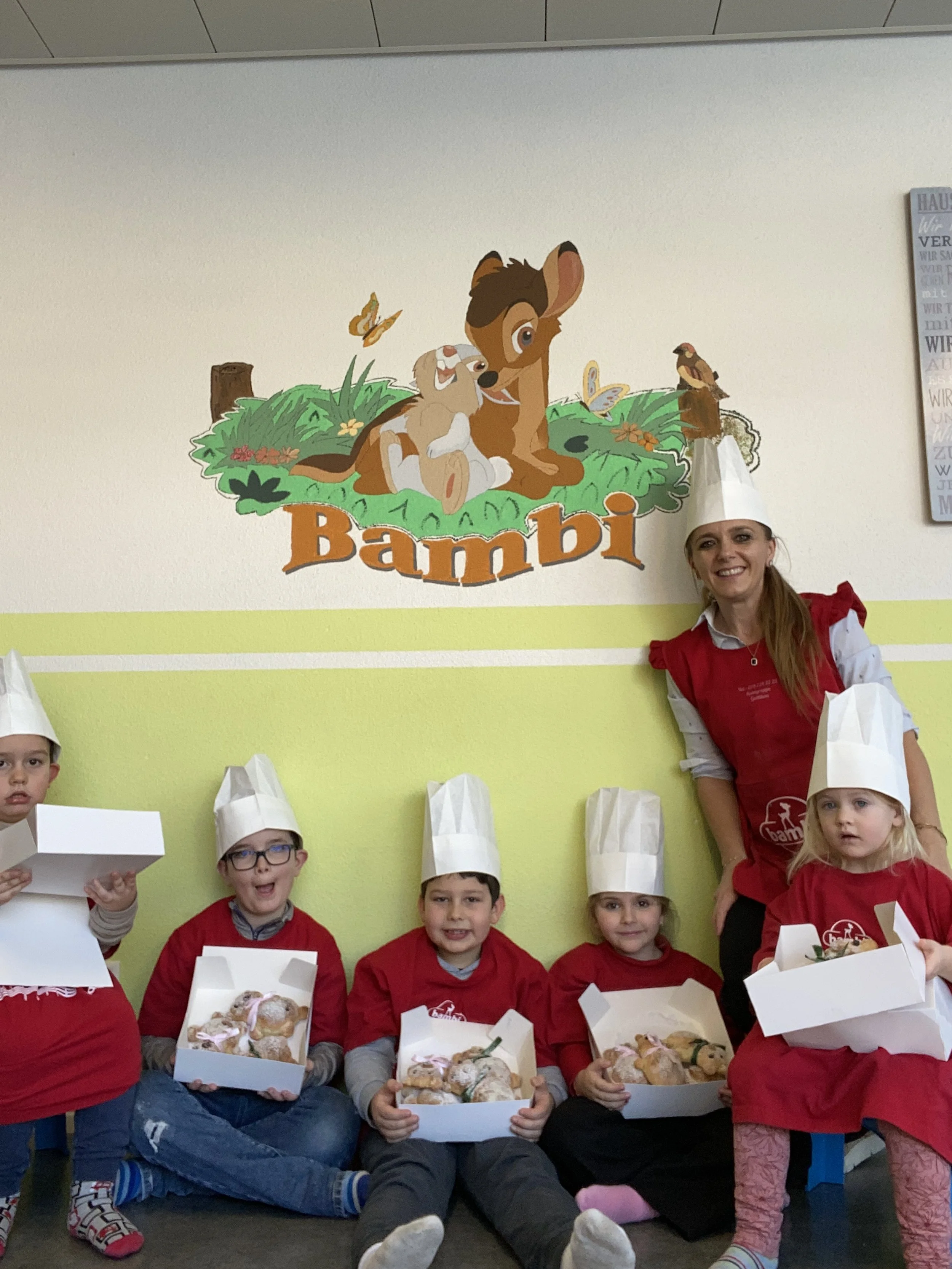 Gruppenfoto von fünf Kindern und einer Erwachsenen Frau in Bambi-Kostümen mit Kochmützen, die Kuchen in Aufbewahrungsboxen halten, vor einer Bambi-Wanddekoration.