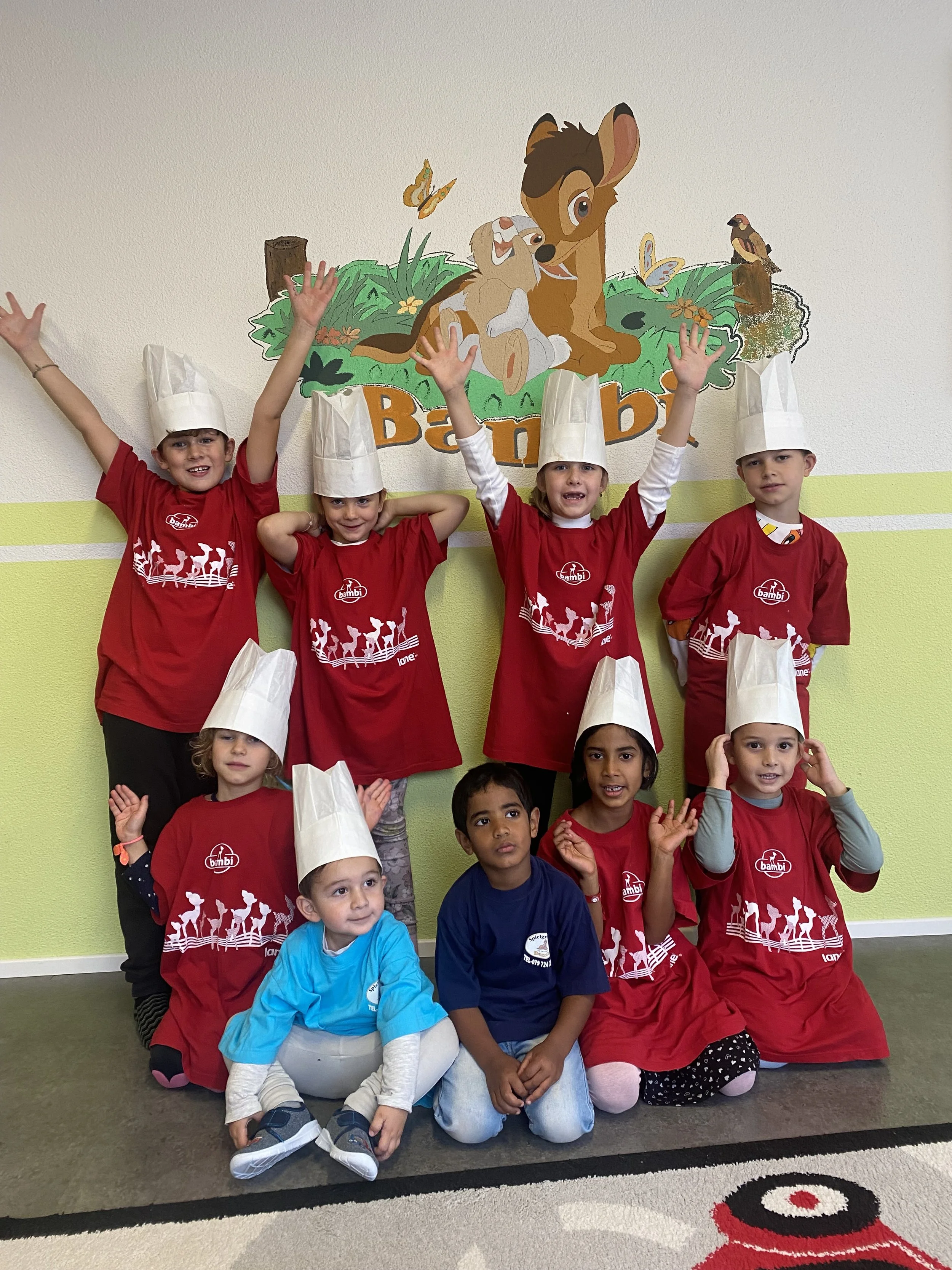 Kinder in Kochmützen und roten T-Shirts, die für ein Kindertheaterstück oder eine Kochveranstaltung posieren, vor einer Wand mit einem Winnie-the-Pooh-Bild und dem Namen 'Bambi'.
