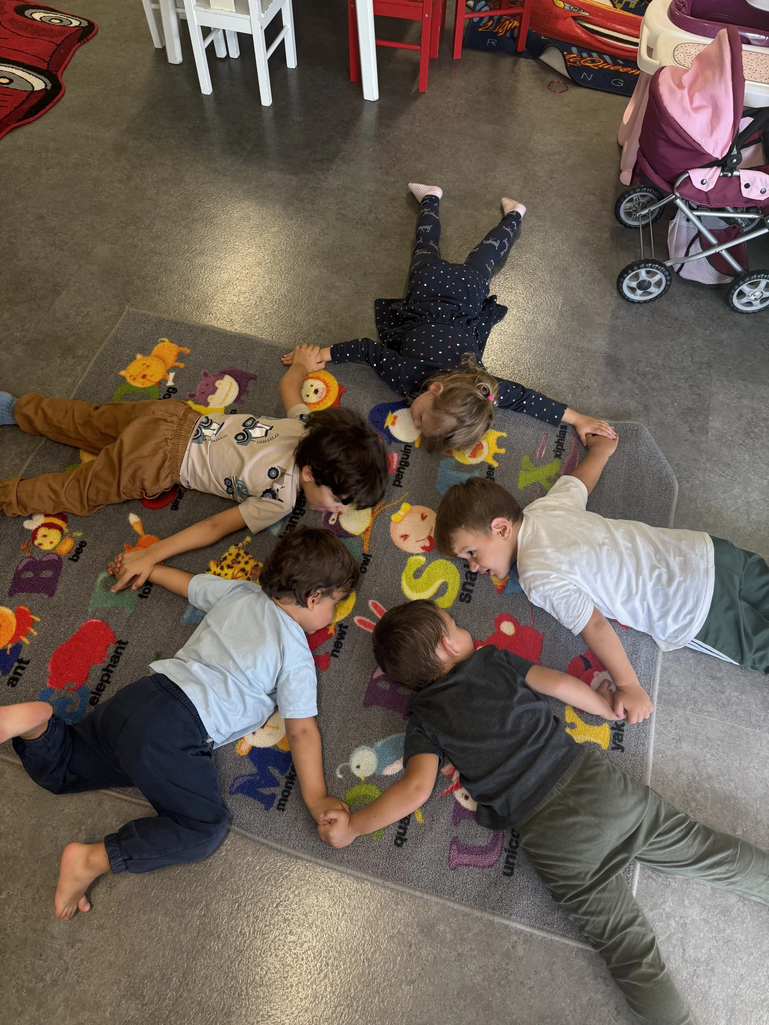 Fünf Kinder liegen in einem Kreis auf einem bunten, tierisch bedruckten Spielteppich im Innenraum, möglicherweise in einem Kindergarten oder Kinderzimmer.