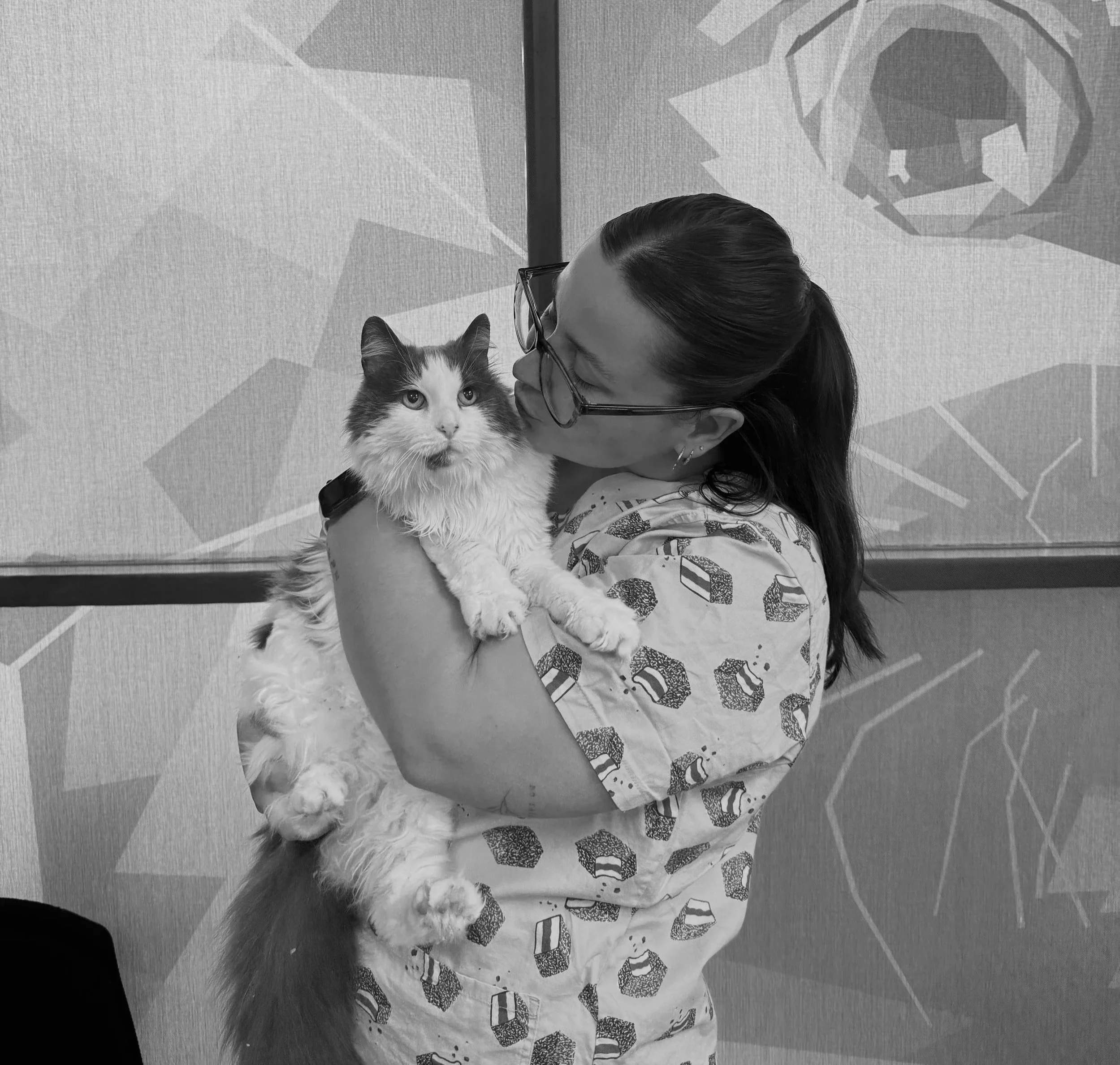 A woman with dark hair wearing glasses and a patterned shirt holds a fluffy, long-haired cat in her arms, leaning in to kiss the cat on its face.