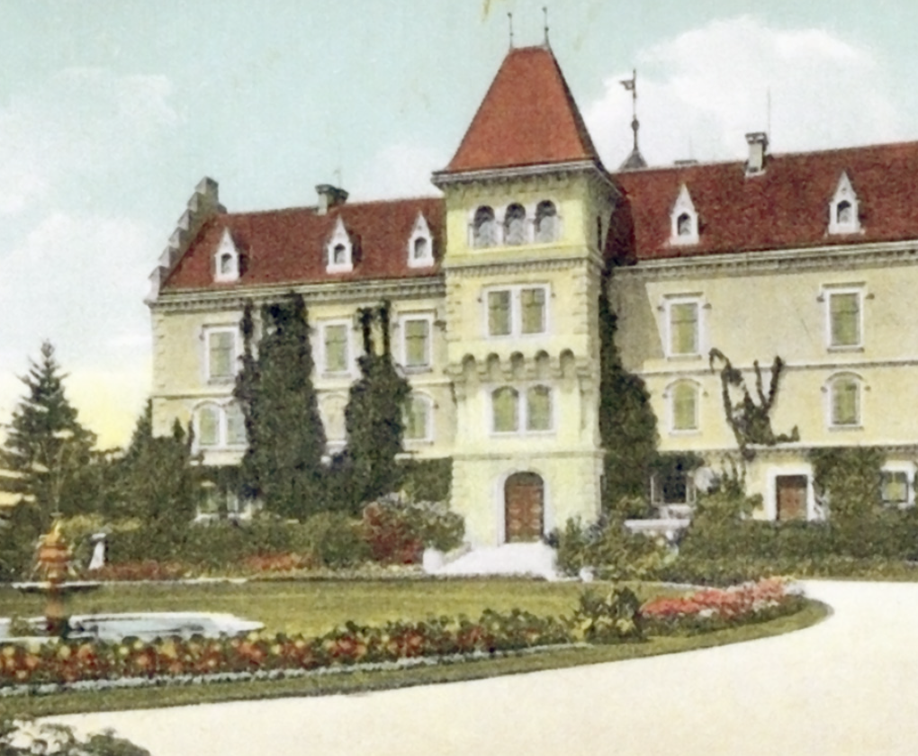 A large historic castle-like building with a red roof, multiple windows, and a central tower. It is surrounded by a garden with colorful flowers and trees.
