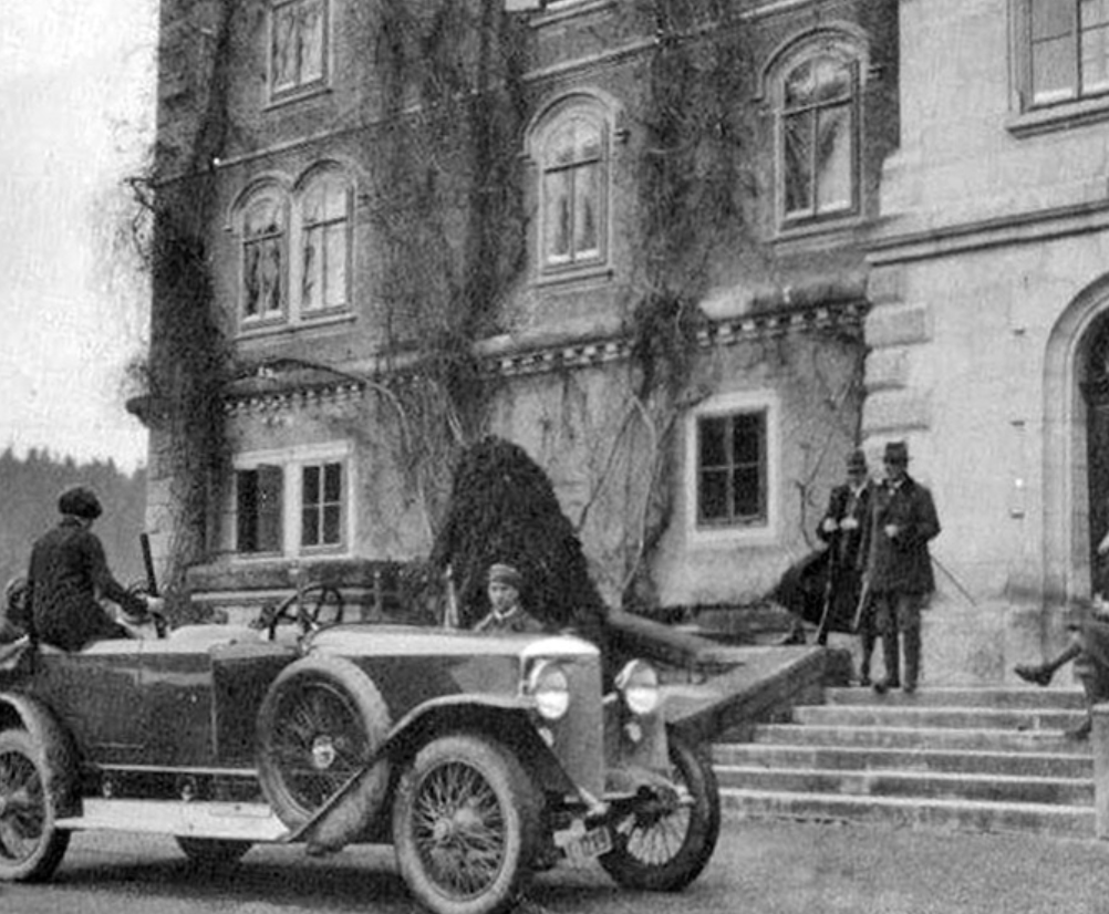 A vintage car parked in front of an old building with four people standing nearby, including two men in suits and hats, a woman, and a child, with a tree growing against the building.