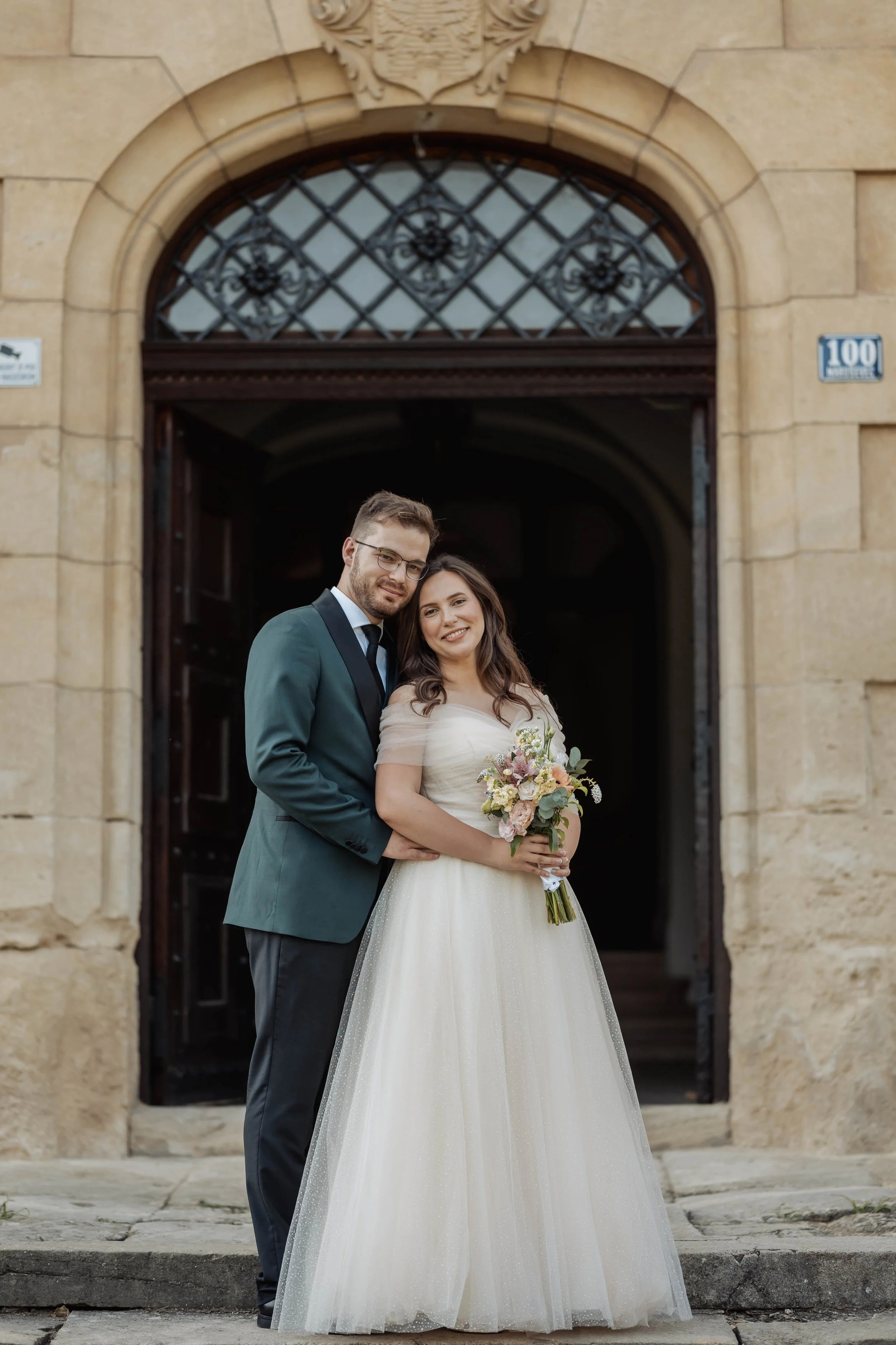 A bride and groom are standing together in front of a grand archway with ornate ironwork above the entrance. The groom is dressed in a blue suit with a black lapel and tie, and the bride is in a white wedding gown holding a bouquet of flowers. They are smiling and close to each other.