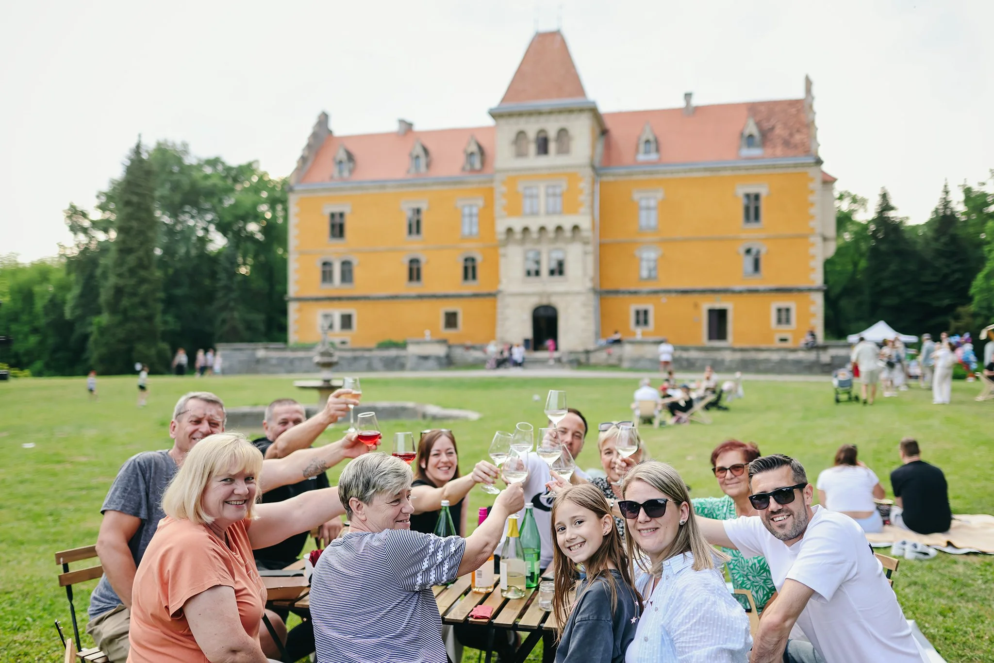 Group of people smiling and raising glasses in a toast outdoors on a lawn in front of a large yellow castle with a red roof, surrounded by trees, on a sunny day.