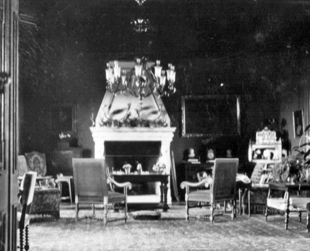 Black and white photo of an ornate living room with a large fireplace at the center, surrounded by chairs and tables, with a chandelier hanging from the ceiling.
