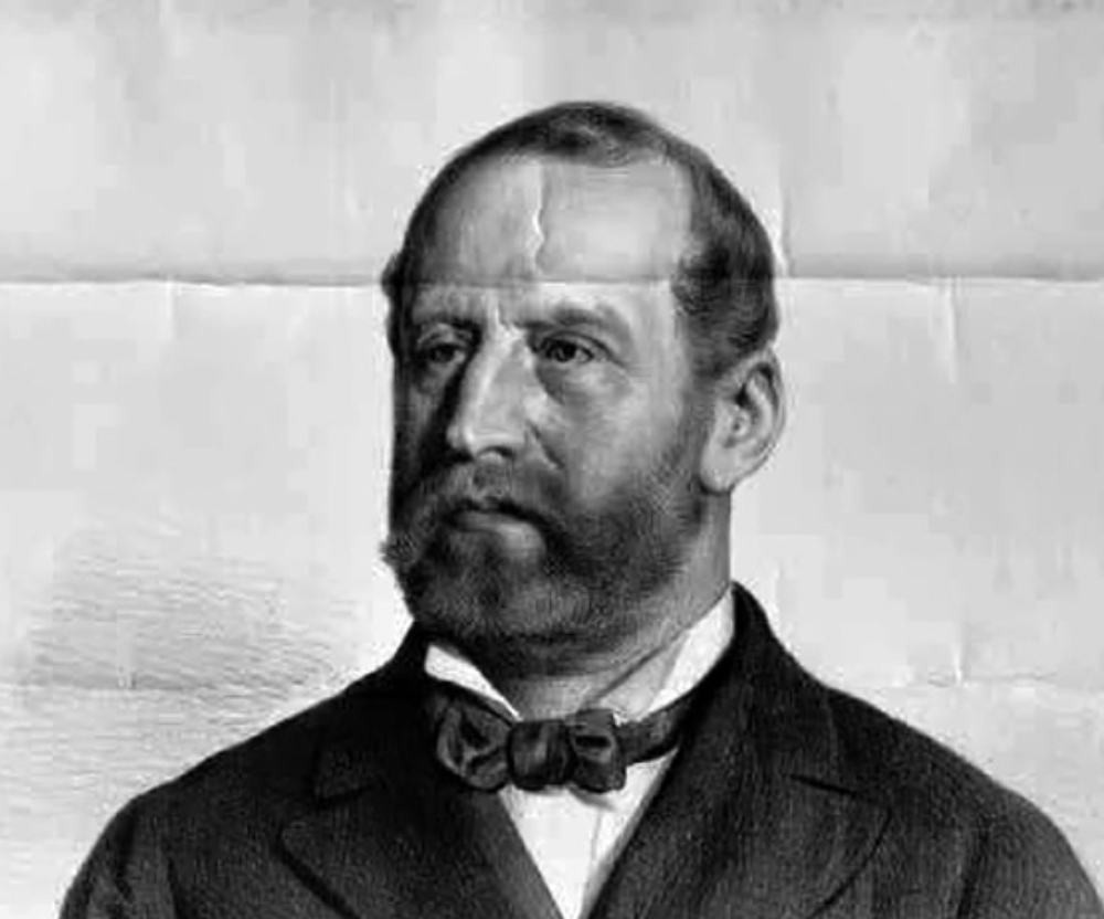 A black-and-white photograph of a man with a beard and mustache, wearing a suit, white shirt, and bow tie, with a serious expression.