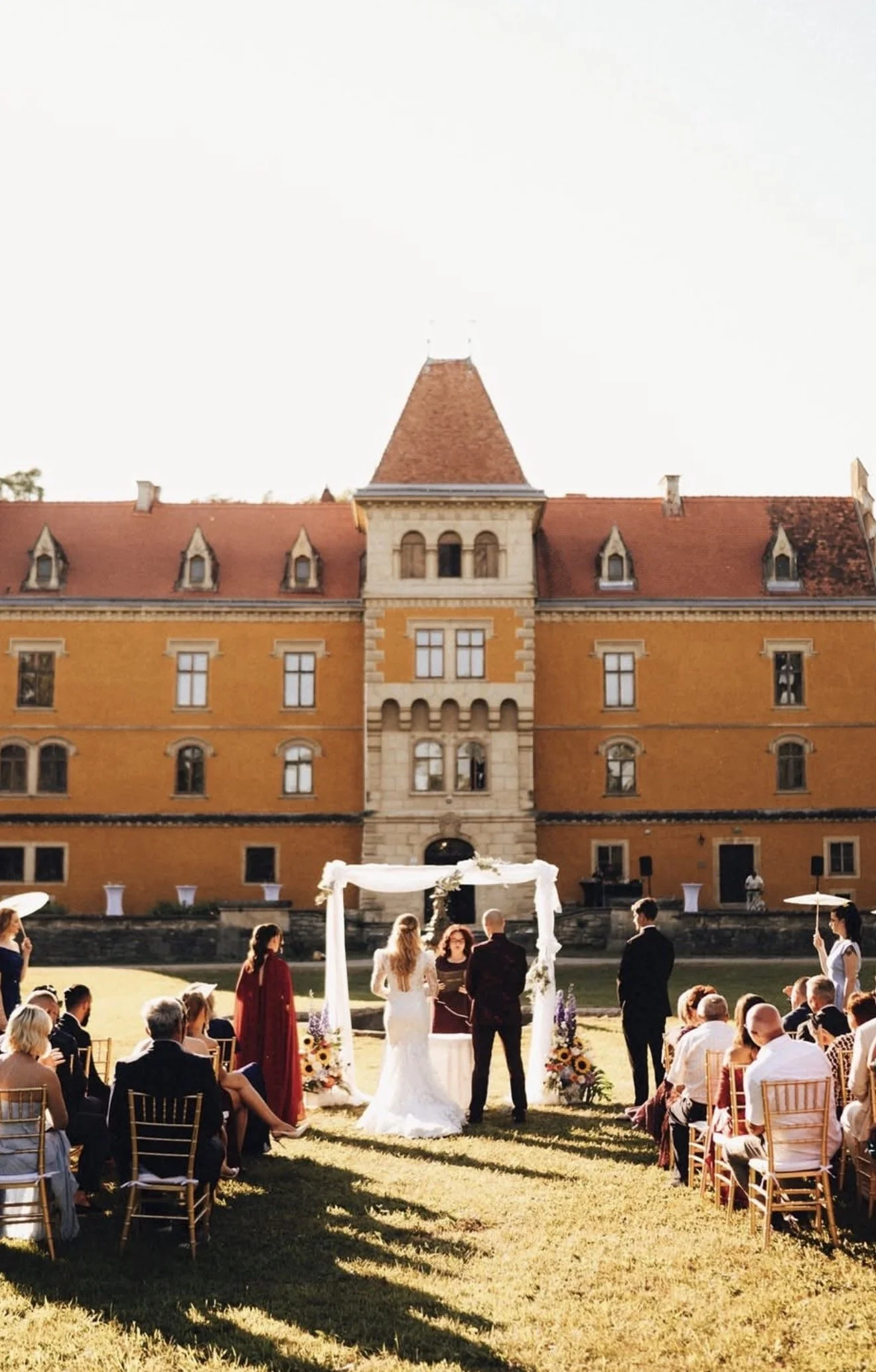 A wedding ceremony taking place outdoors in front of a historic brick building with a high pointed roof, windows, and a central entrance. The bride and groom are standing under a white arch decorated with flowers, surrounded by friends and family seated on chairs, with sunlight casting long shadows on the grass.