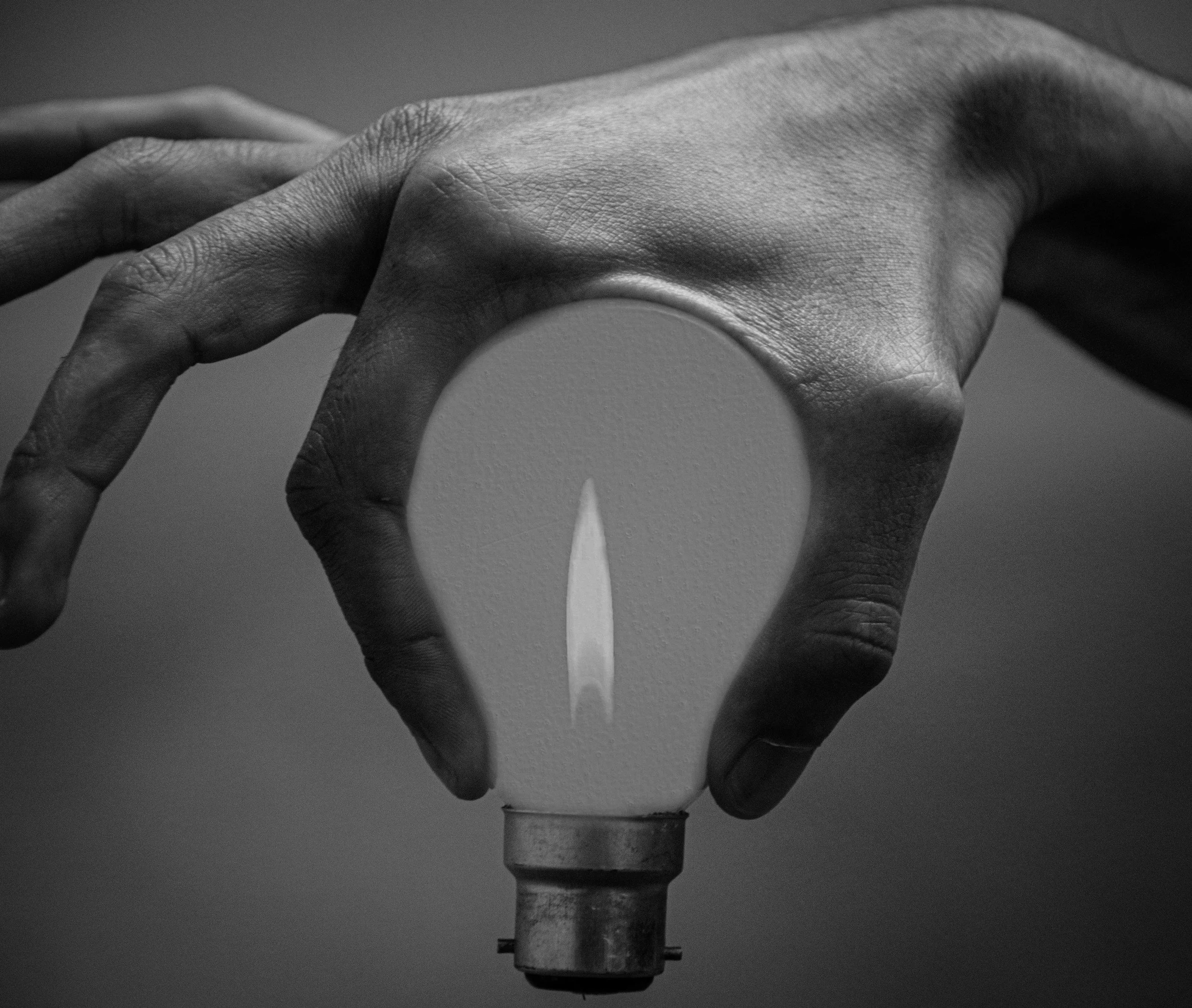 A person's hand holding a broken light bulb with a visible flame inside, in black and white.