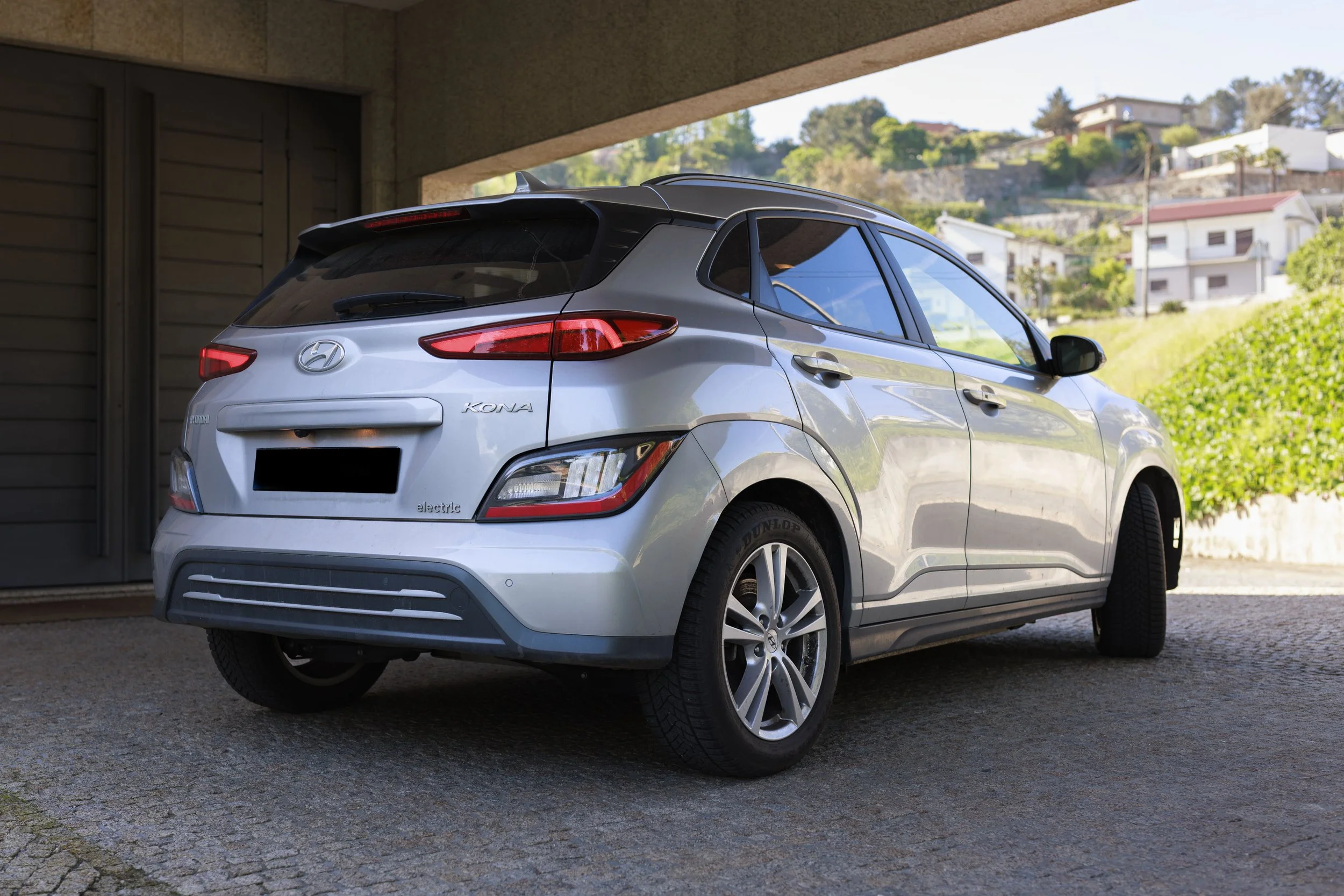 Carro hatchback cinza estacionado em uma garagem com fundo de casas e morros verdes.