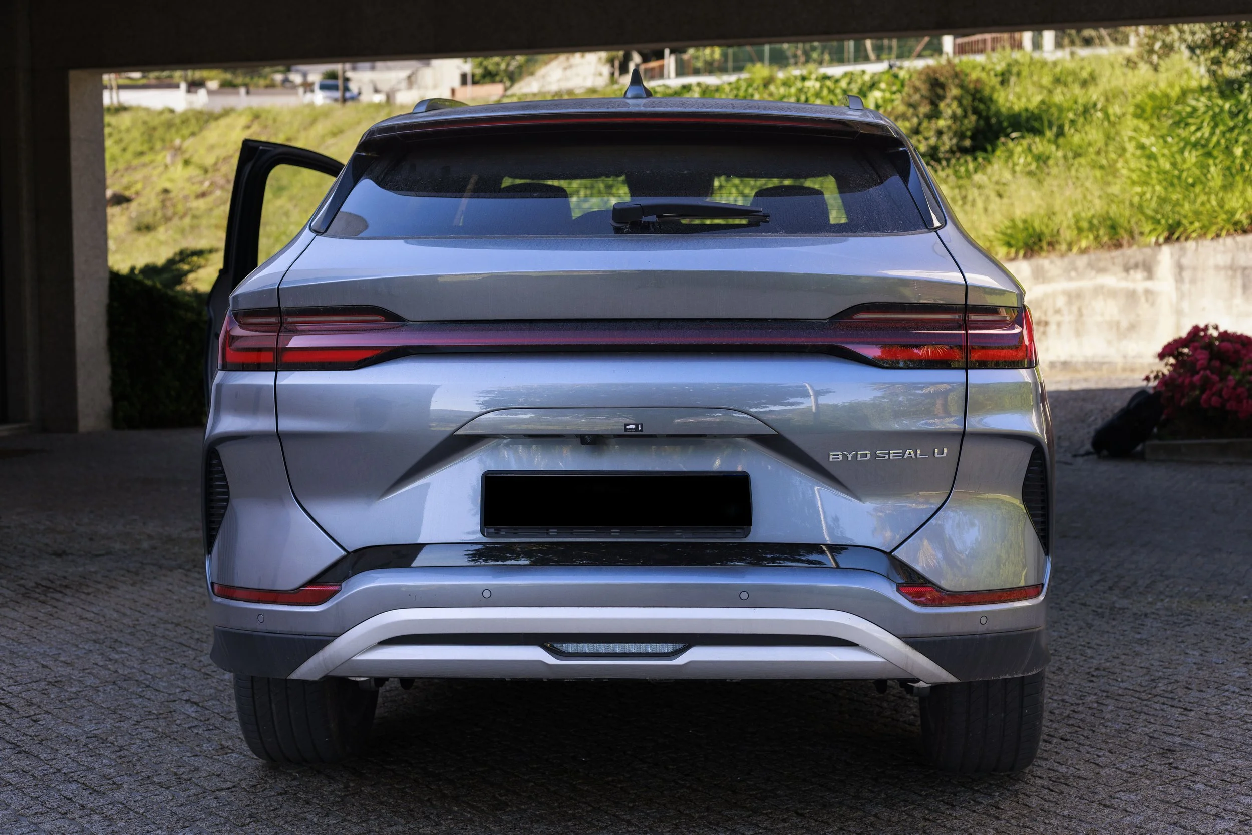 Carro SUV prata estacionado ao ar livre, com porta aberta na lateral esquerda, sob uma estrutura de sombra, com vegetação e um muro ao fundo.