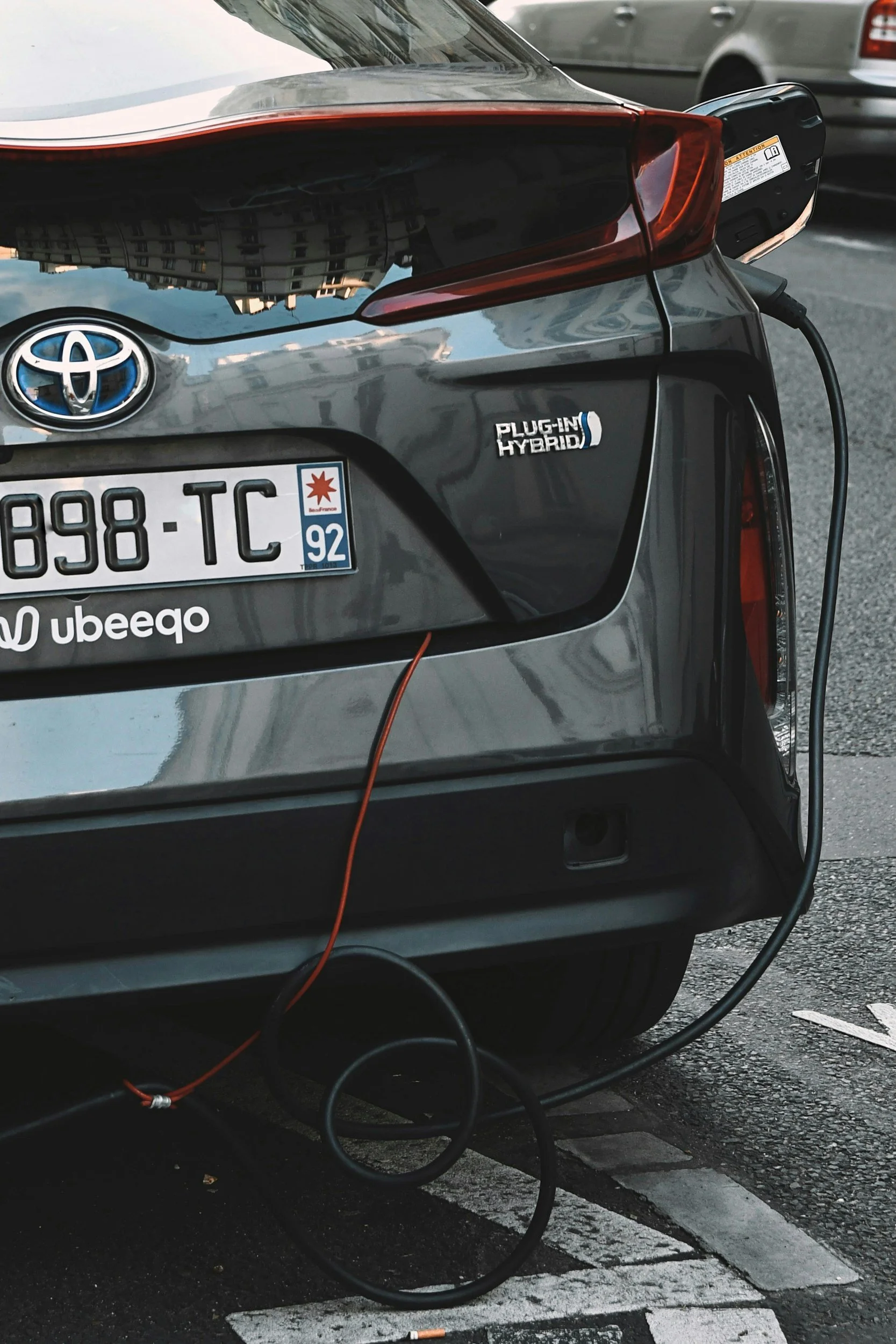 O retro do carro de cor cinza mostra o emblema da Toyota e um adesivo de carro híbrido plug-in. Há um carregador conectado na traseira do veículo, com cabos de carregamento na rua, indicando que o carro elétrico está sendo carregado.