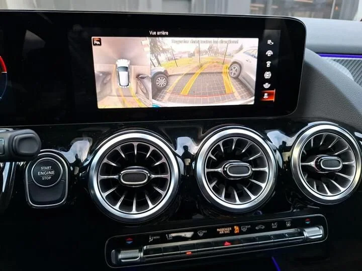 Tela de monitoramento de estacionamento exibindo uma visualização de câmera de ré com linhas guia amarelas, ao lado de dois dois círculos de ventilação no painel do carro.