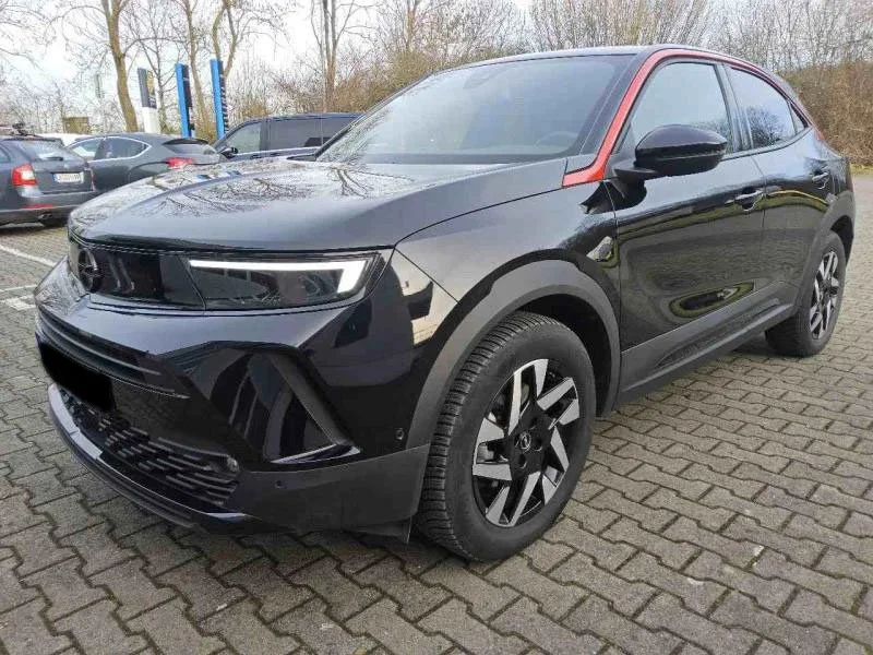Carro hatchback preto estacionado em uma área de estacionamento de paralelepípedo, com árvores ao fundo.