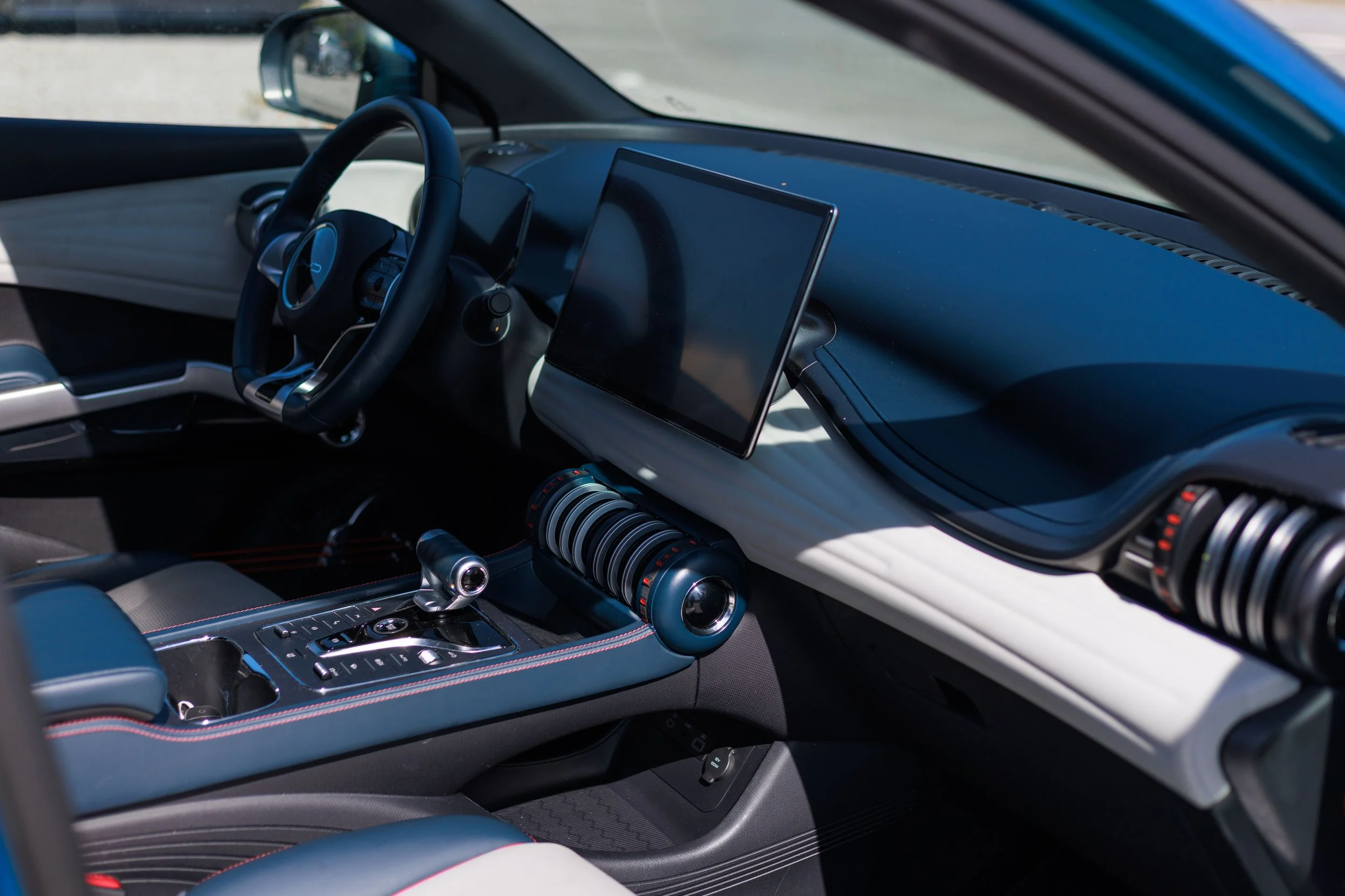 Interior de um carro futurista com painel digital, volante esportivo, e pôsteres digitais, com sedes revestidas em couro azul com costura vermelha.