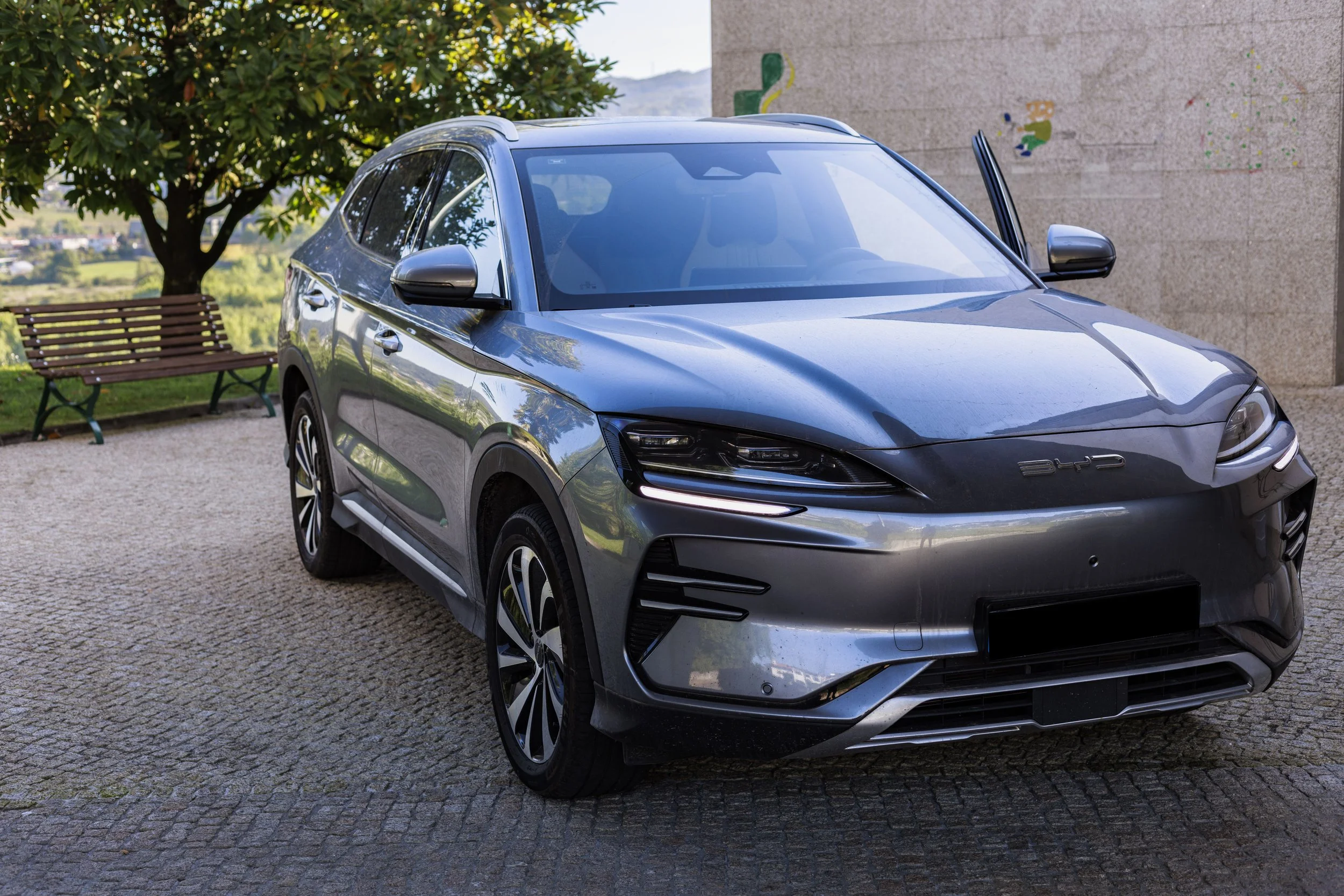 Carro de cor cinza estacionado em recanto com bancos de madeira e árvore ao fundo.