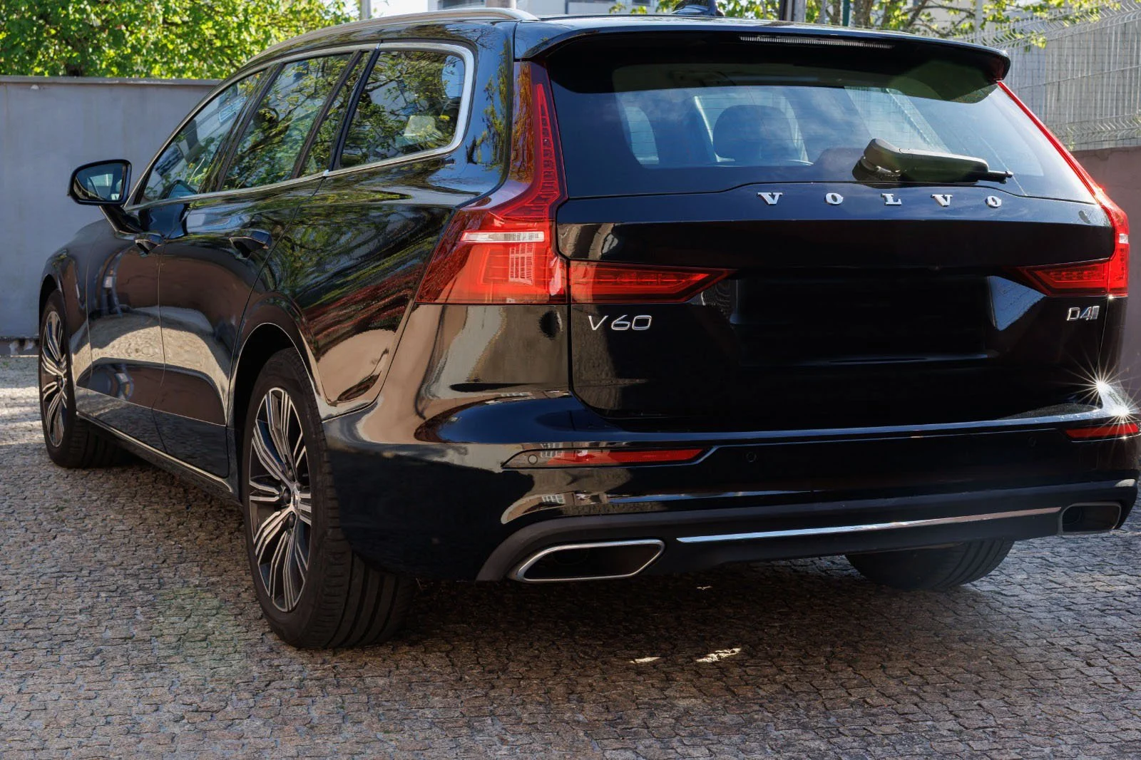 Carro preto da marca Volvo, modelo V60, visto de trás, com embalagem de vidro e detalhes em prata, estacionado em uma rua de paralelepípedos, com fundo de muro branco e árvores ao redor.