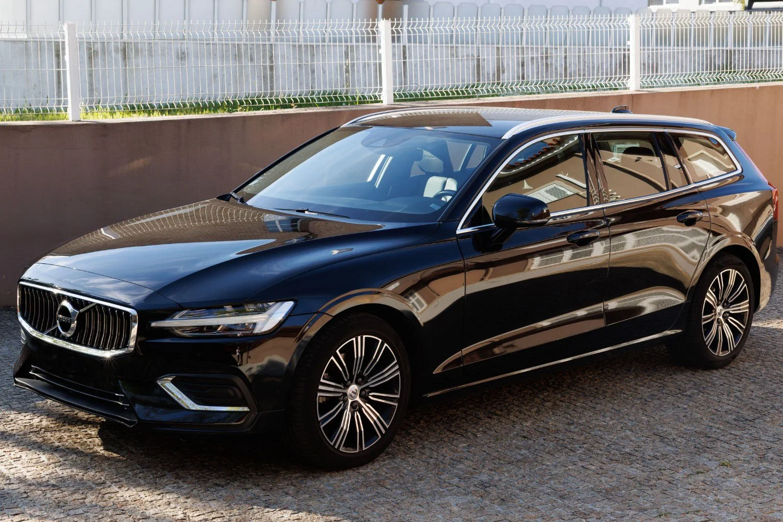 Carro preto do modelo sedan com design moderno, com detalhes em prata, estacionado na calçada ao lado de uma cerca branca