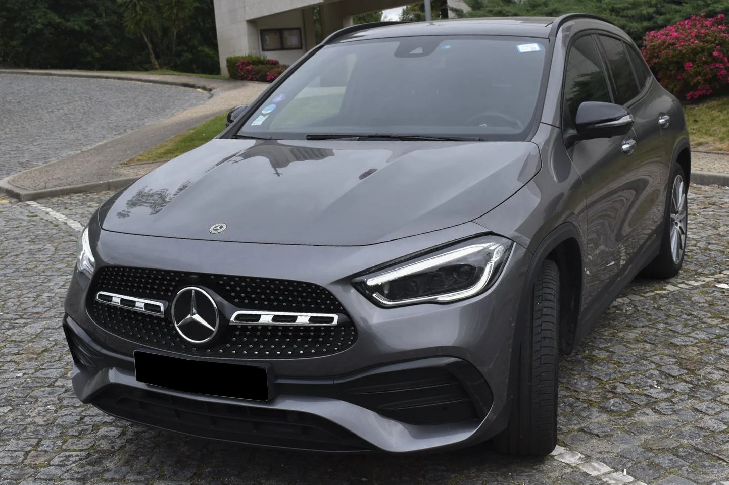 Carro cinza do modelo Mercedes-Benz em estacionamento de rua com piso de pedra e vegetação ao fundo.