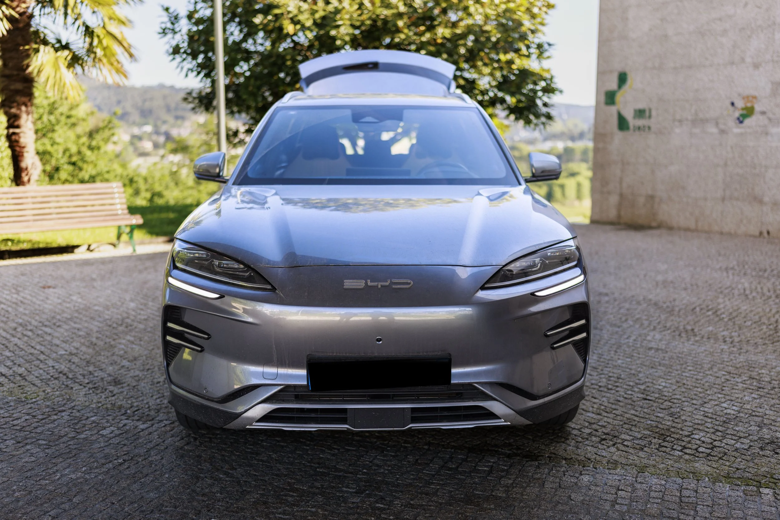 Carro elétrico prata estacionado em uma calçada de pedra, com árvores e edifícios ao fundo.