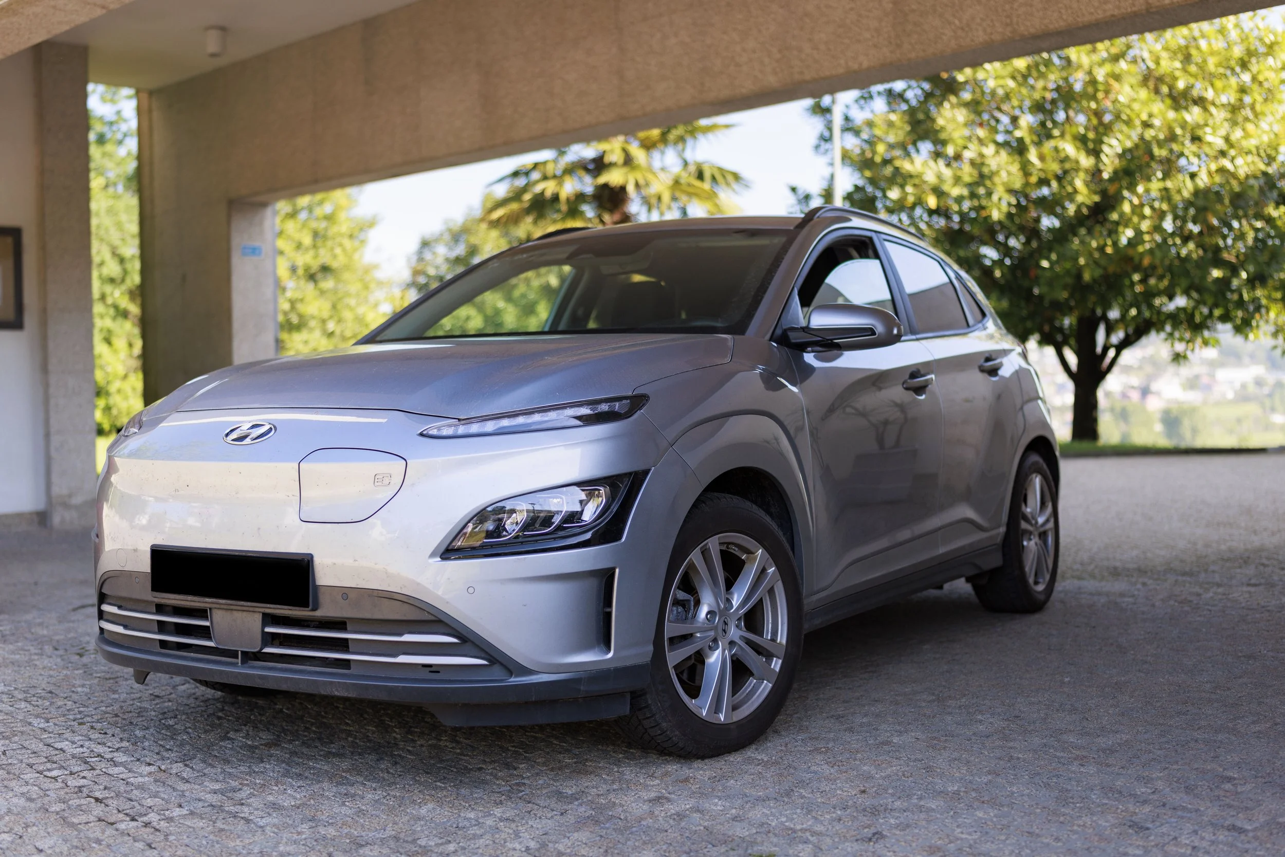 Carro cinza de modelo hatchback estacionado sob marquise com árvores verdes ao fundo.