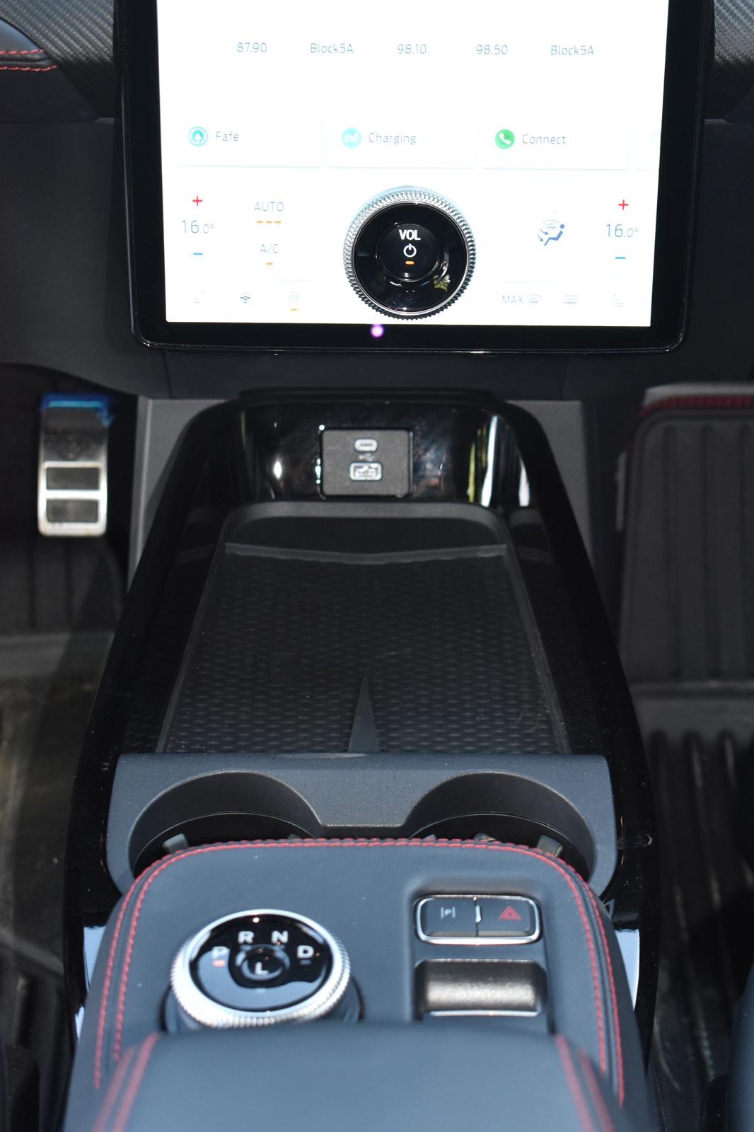 Painel de controle do carro com tela touchscreen, controles de volume, temperatura, e botão de câmbio giratório. À esquerda, pedal de freio, e ao centro, espaço para objetos.