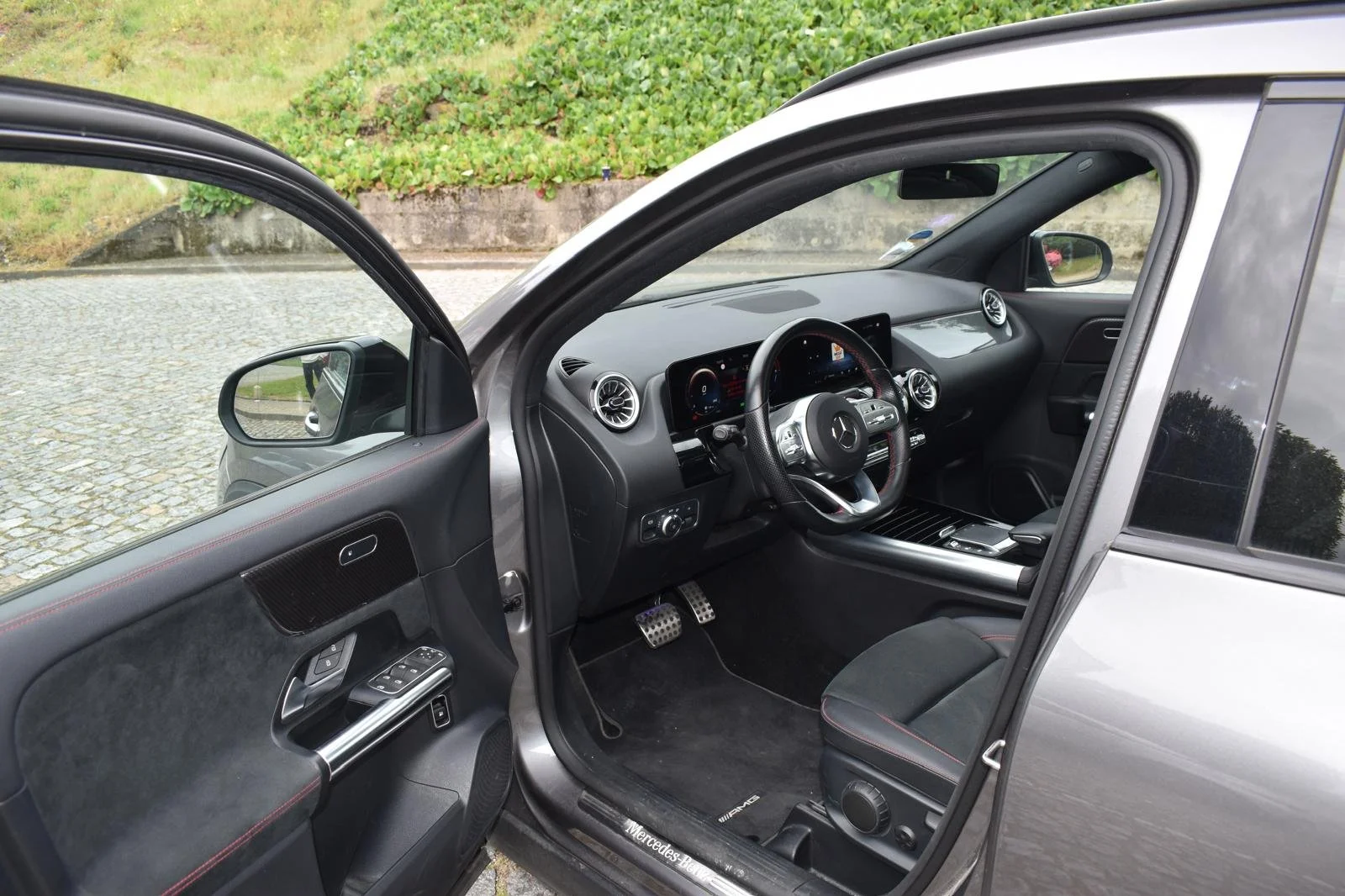 Interior de um carro Mercedes-Benz, focando no painel, volante digital, assentos de couro esportivos e pedais de metal com borracha antiderrapante, visível pelo lado do motorista com as portas abertas.