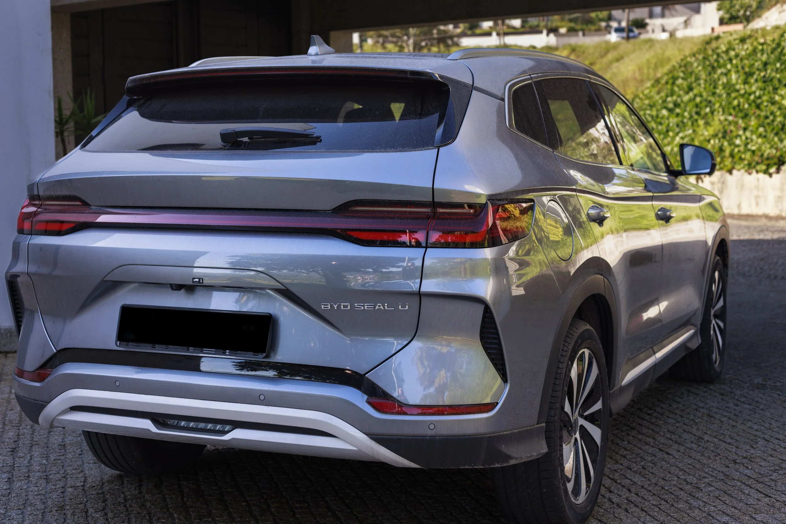 Vista traseira de um carro prata de luxo, provavelmente um SUV, estacionado em uma área pavimentada ao ar livre, com vegetação verde ao fundo.