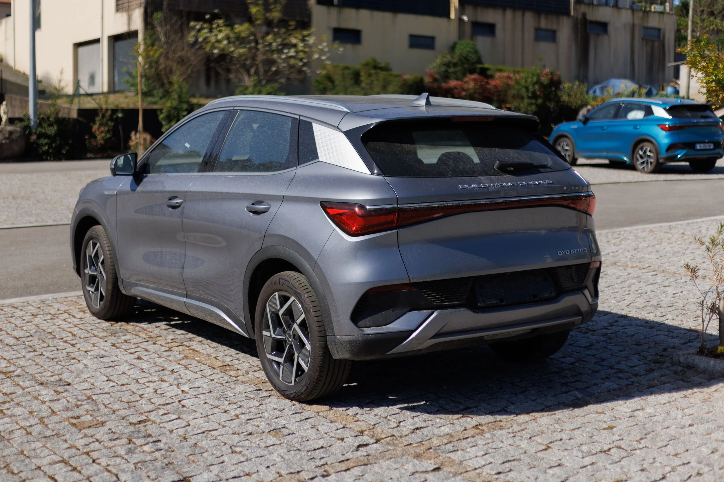 Carro SUV cinza estacionado na rua de pedra, com outros carros azuis ao fundo, em frente a edifícios residenciais cercados de árvores e arbustos.