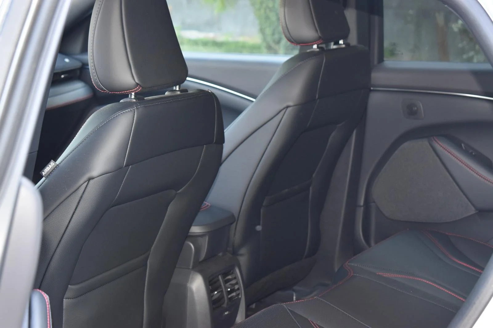 Interior de um carro visto de trás. Assentos pretos com costura vermelha, painel e porta dianteiros visíveis.