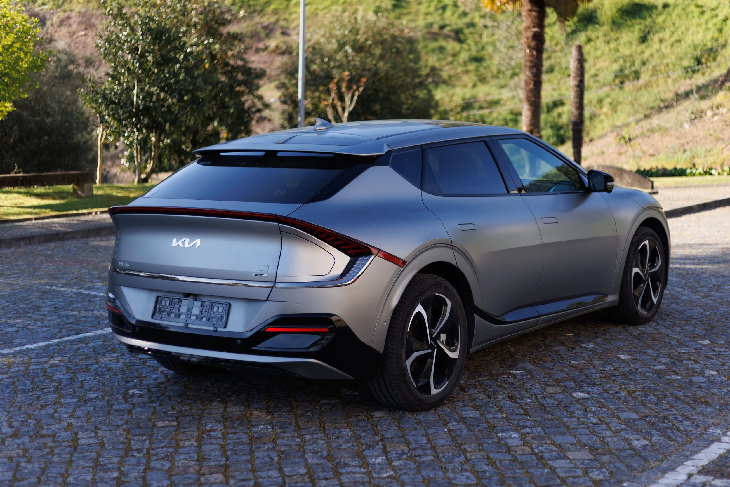 Carro cinza futurista estacionado em avenida com calçada de pedra, árvores e vegetação ao fundo.