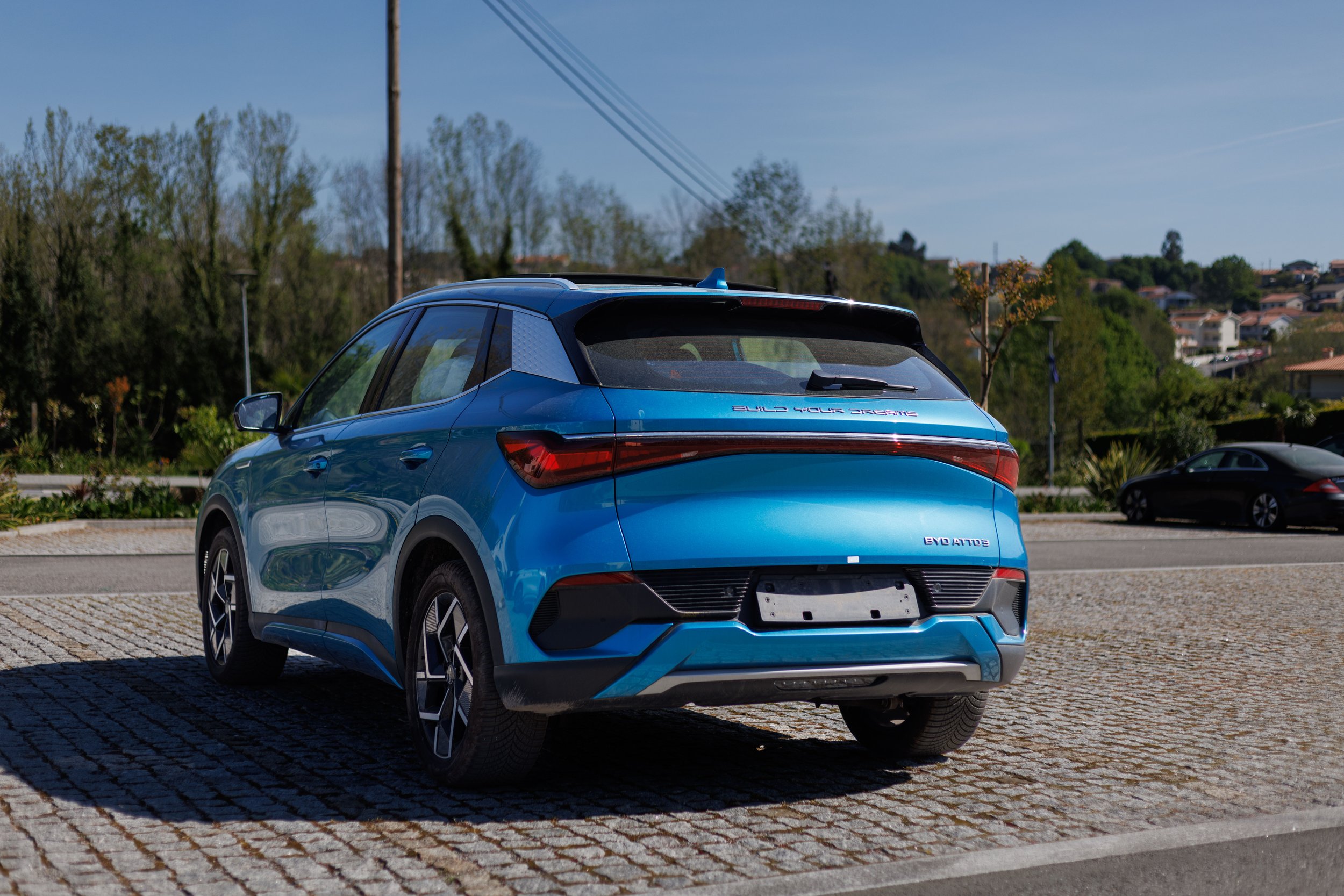 Veículo elétrico azul estacionado em uma praça com árvores e casas ao fundo.