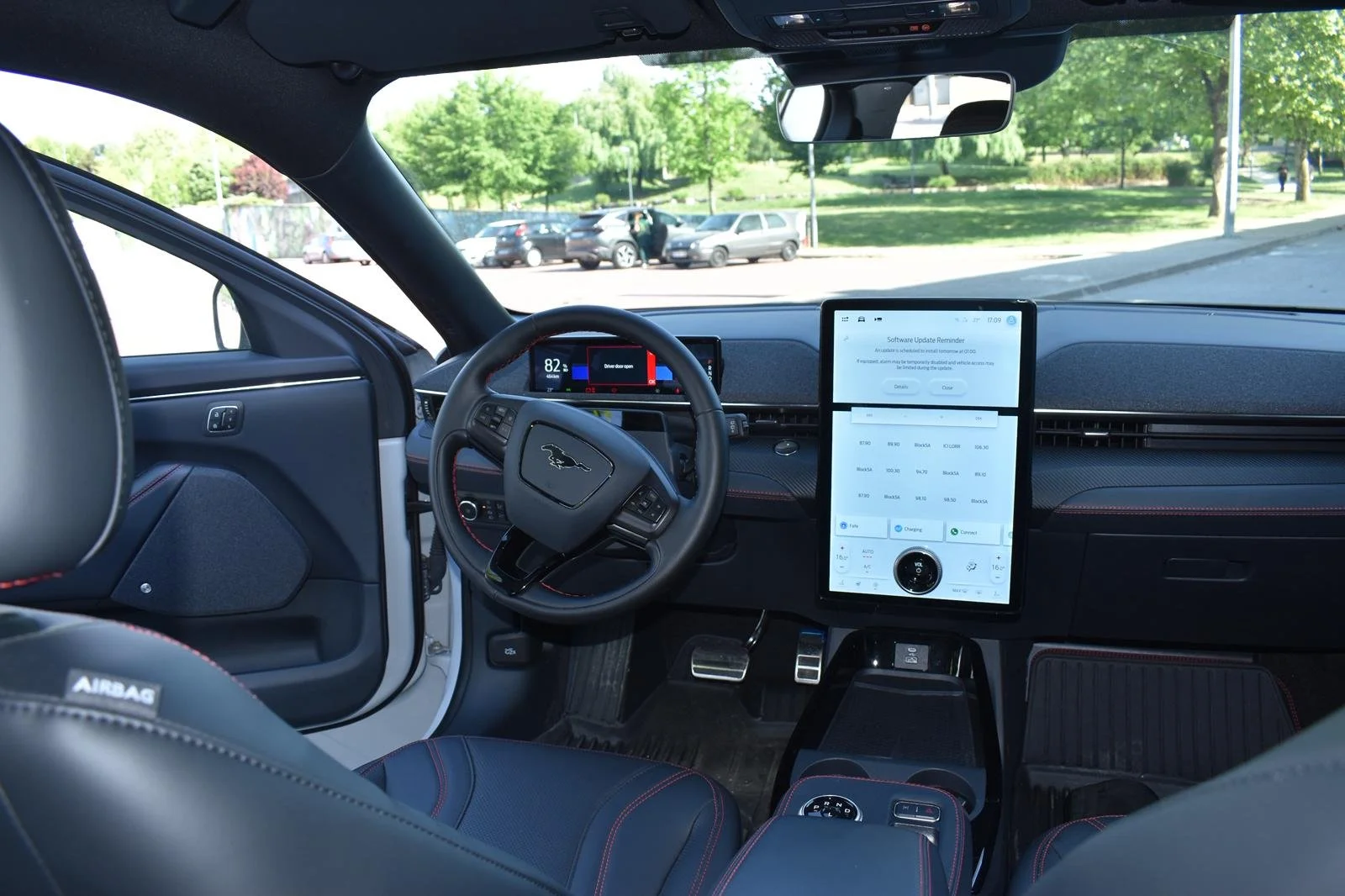 Interior de um carro elétrico com painel digital, volante com logo de um guepardo, tablet grande na direção exibindo detalhes do sistema do veículo, assento de couro com costura vermelha, árvores visíveis pela frente e janela aberta.